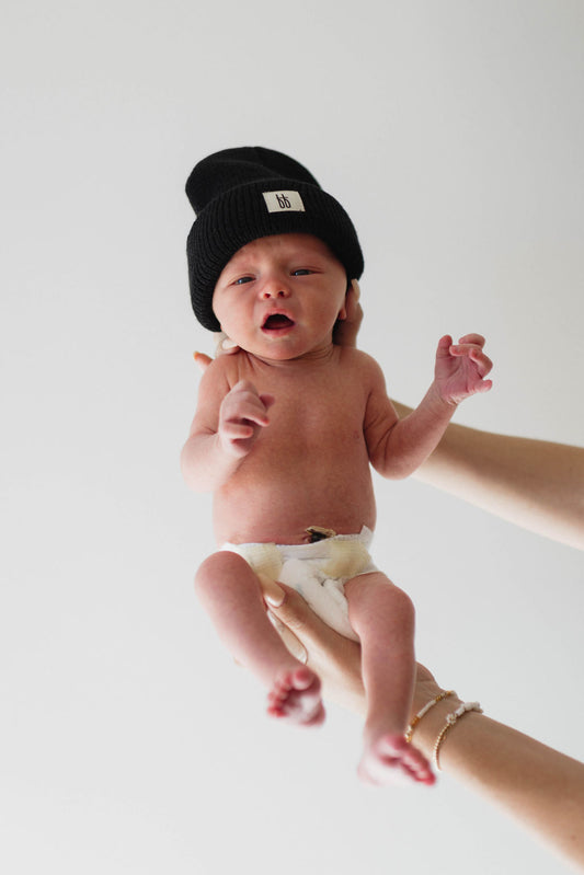 A newborn baby wearing the ff Knit Beanie in Black by forever french baby, paired with just a diaper, is being held up gently by adult hands. The baby has a calm expression and small, bundled fists against a white background, creating a serene atmosphere. This timeless moment captures the essence of forever french baby.