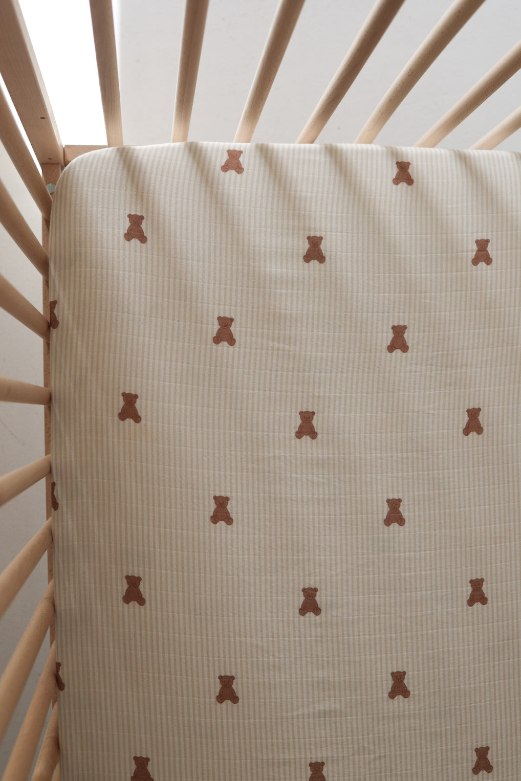 The forever french baby Muslin Crib Sheet | Teddy features a cute brown teddy bear design on subtle stripes, shown here on a light wood crib. Soft light enhances the cozy atmosphere—perfect for your baby's bedding.