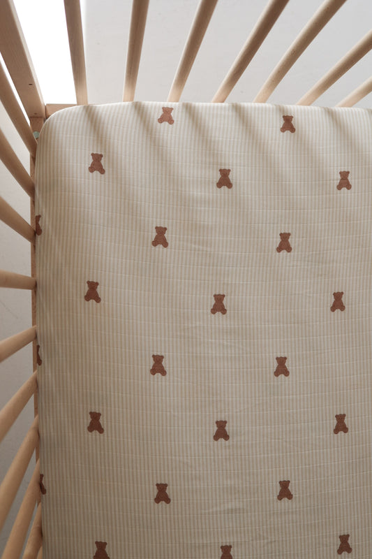 The forever french baby Muslin Crib Sheet | Teddy features a cute brown teddy bear design on subtle stripes, shown here on a light wood crib. Soft light enhances the cozy atmosphere—perfect for your baby's bedding.
