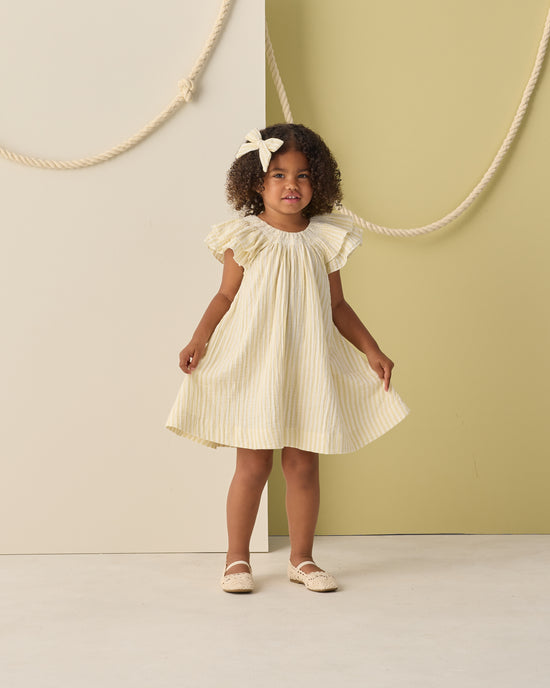 A young girl with curly hair, wearing the Rylee + Cru Lucia Dress in Citron Stripe—a yellow and white mini dress paired with a matching hair bow—smiles as she stands against a neutral background accented with rope decorations.
