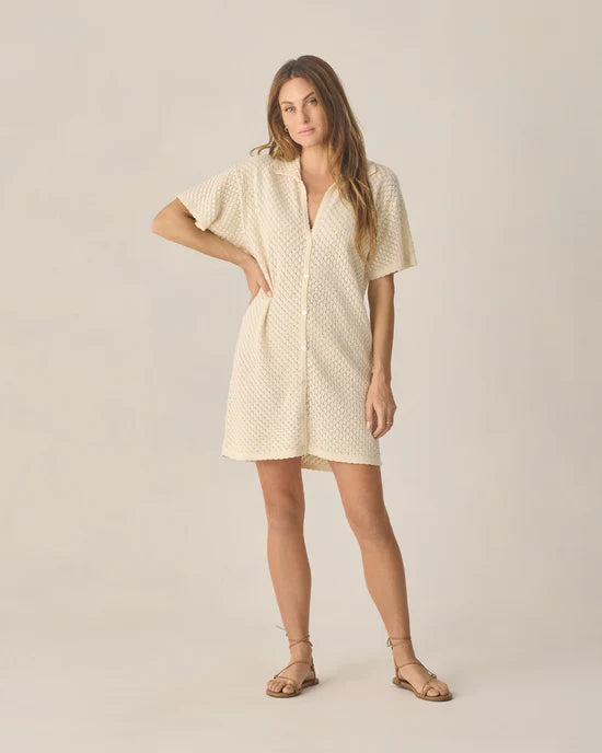 A woman stands wearing the Rylee + Cru Women’s Crochet Cover Up in Natural—a short, cream-colored, textured button-up dress featuring a flattering v-neckline and short sleeves. Made from 100% cotton, this cover up pairs effortlessly with her flat sandals. She has long hair, and the background is plain and light-colored.