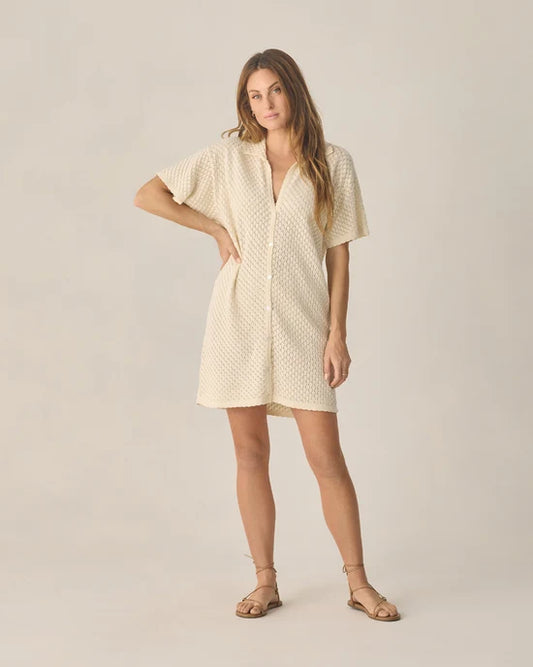 A woman stands wearing the Rylee + Cru Women’s Crochet Cover Up in Natural—a short, cream-colored, textured button-up dress featuring a flattering v-neckline and short sleeves. Made from 100% cotton, this cover up pairs effortlessly with her flat sandals. She has long hair, and the background is plain and light-colored.