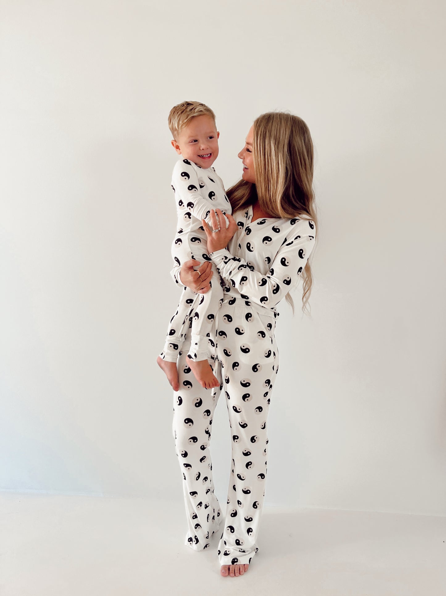 A woman with long blonde hair is holding a smiling young boy. Both are wearing matching Women's Bamboo Pajamas | Black & White Yin Yang from forever French baby. They stand against a plain light-colored backdrop, looking at each other with cheerful expressions in their breastfeeding-friendly outfits.