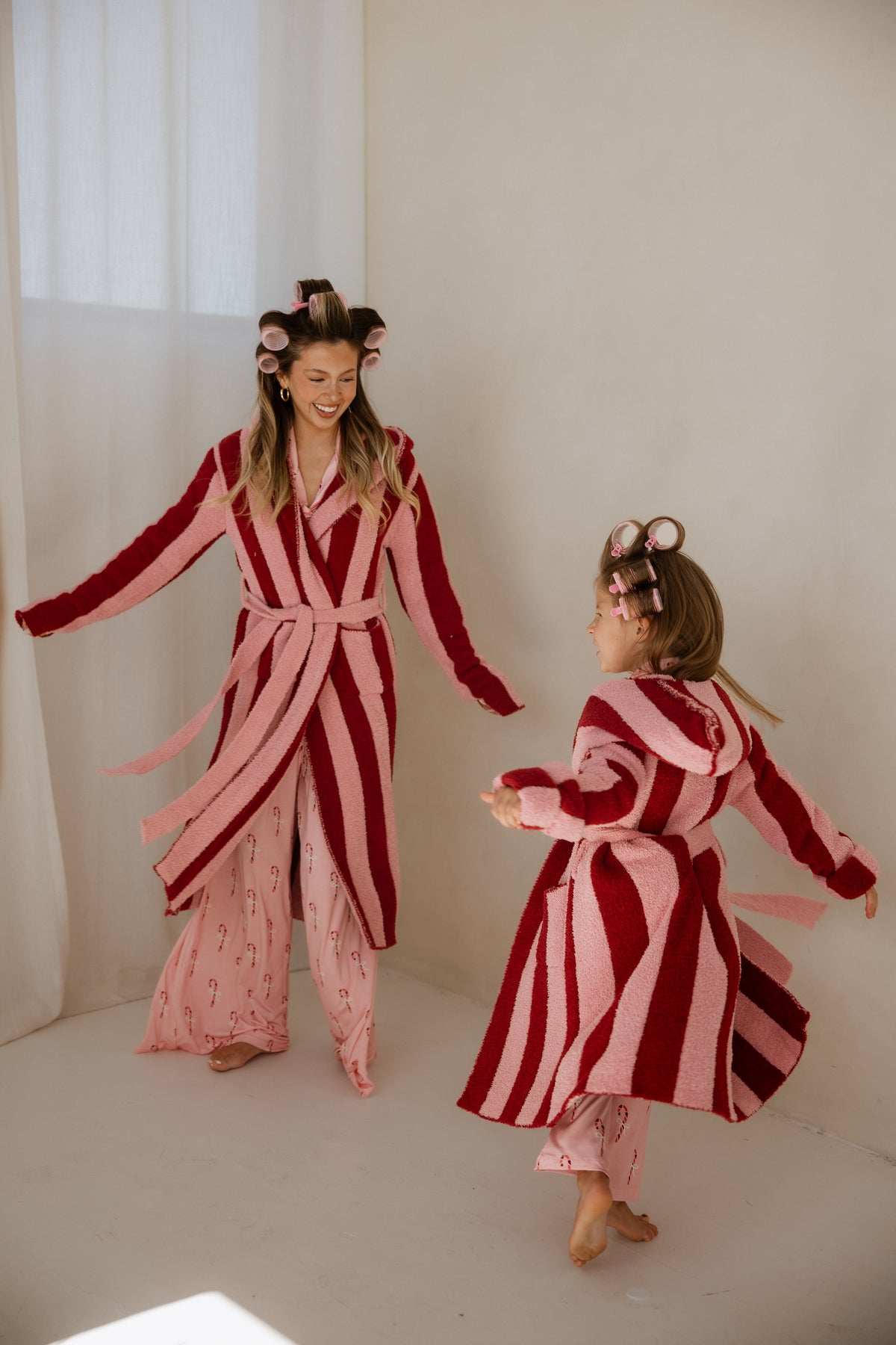 A woman and a young girl wearing matching pink and dark red Sugar Plum Stripe hooded robes and hair rollers, twirling and smiling.
