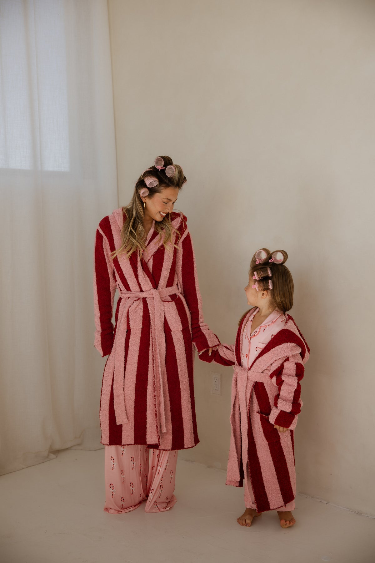 A woman and a young girl wearing matching Adult Robe | Sugar Plum Stripe robes in pink and red stripes and hair rollers hold hands.