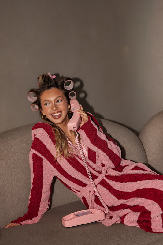 A person with hair rollers in a pink and red striped Adult Robe | Sugar Plum Stripe reclines on a sofa while talking on a vintage pink corded telephone.