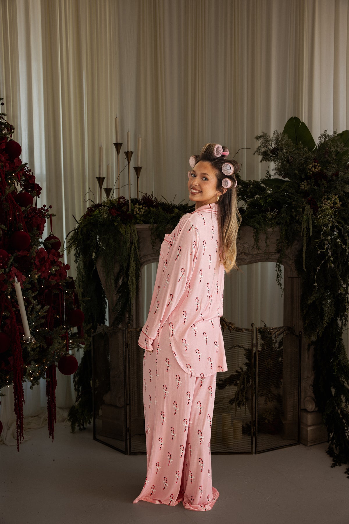 A woman wearing a Women’s Bamboo Button Up Lounge Set | Pink Candy Cane and hair rollers standing in front of a fireplace and Christmas tree.