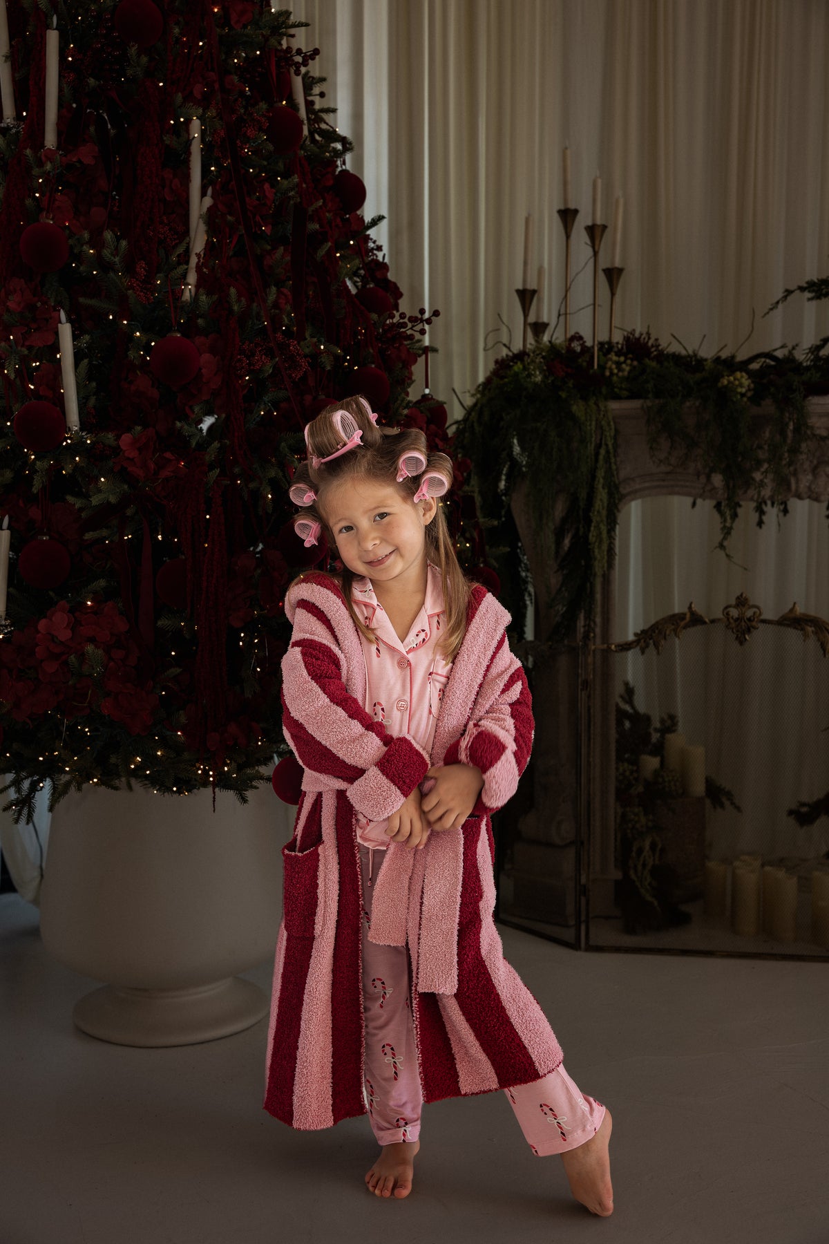 A child in pink pajamas wears a Children’s Robe | Sugar Plum Stripe made of microfiber feather yarn, standing in front of a decorated Christmas tree.