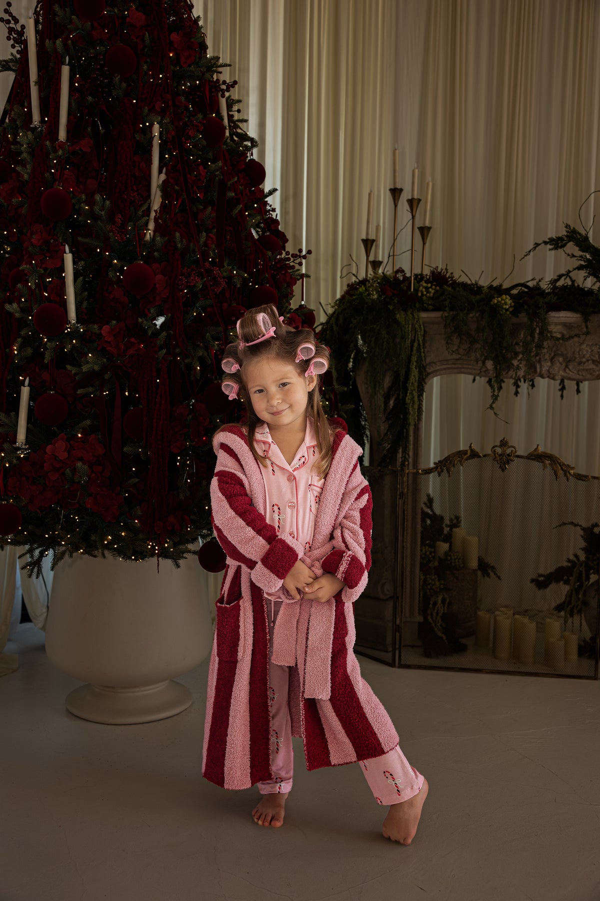 A young girl in hair rollers and a Girl’s Bamboo Button Up Lounge Set | Pink Candy Cane with a matching robe stands in front of a decorated Christmas tree.