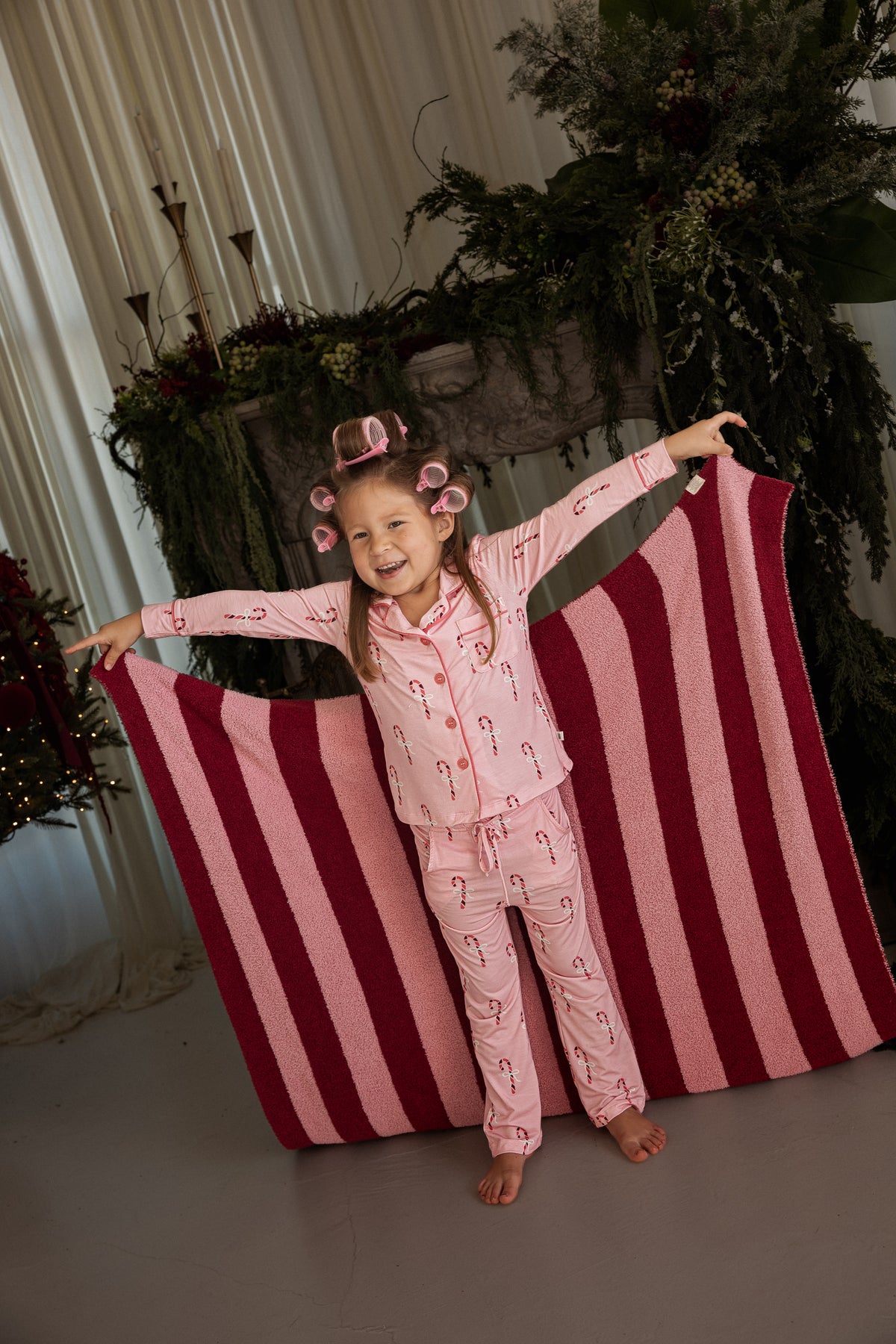 A smiling girl in pink candy cane pajamas and hair rollers holds up a large *PRE-ORDER* Plush Blanket | Sugarplum Stripe.