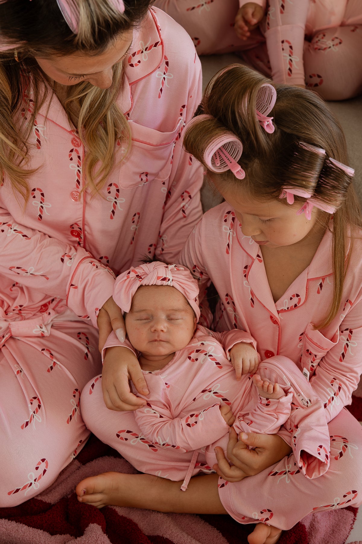 A woman and a young girl wearing matching Bamboo Zip Pajamas | Pink Candy Cane hold a newborn baby in a matching outfit and headband.