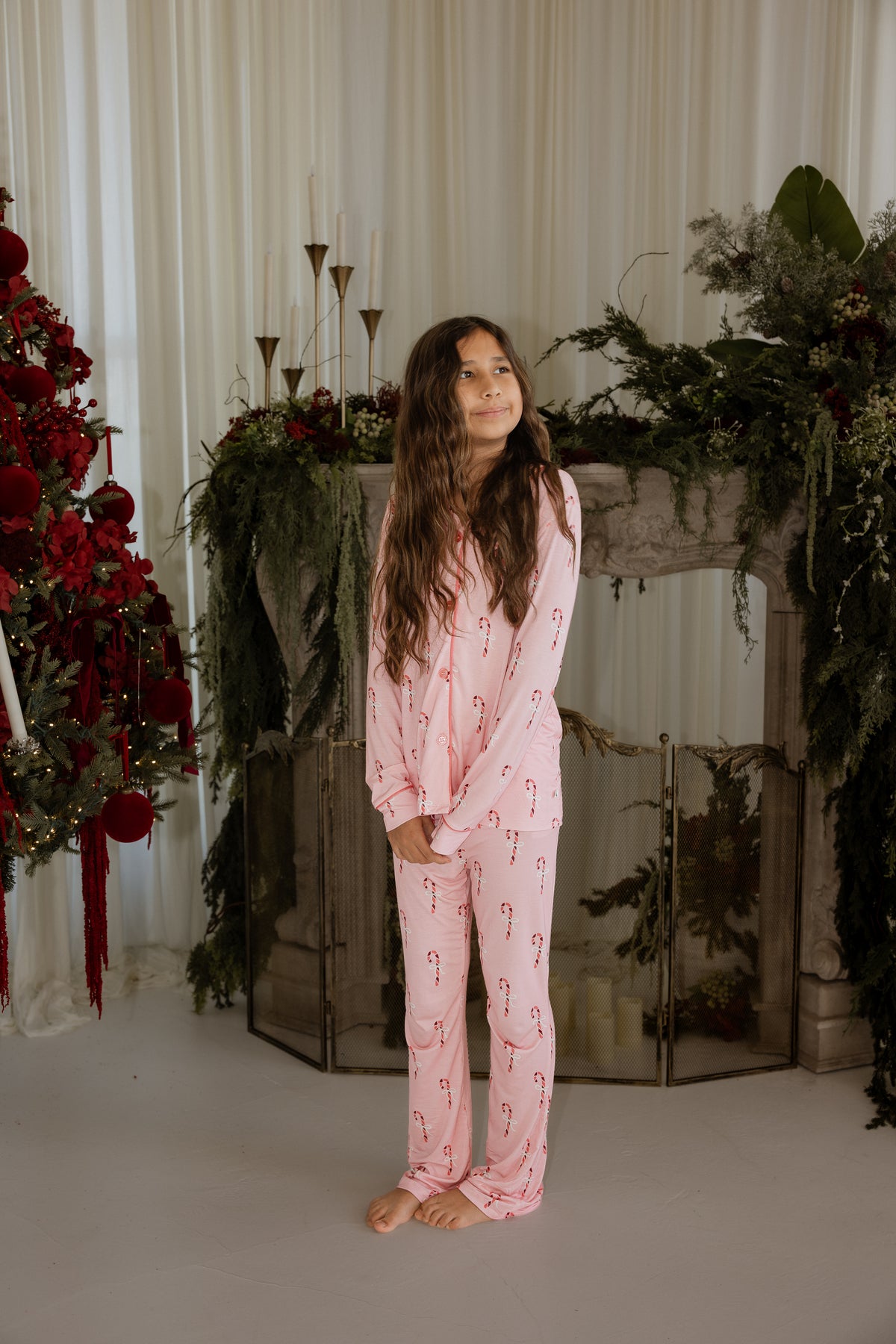 A girl stands in front of a fireplace wearing a light pink Girl’s Bamboo Button Up Lounge Set | Pink Candy Cane patterned with small candy canes.
