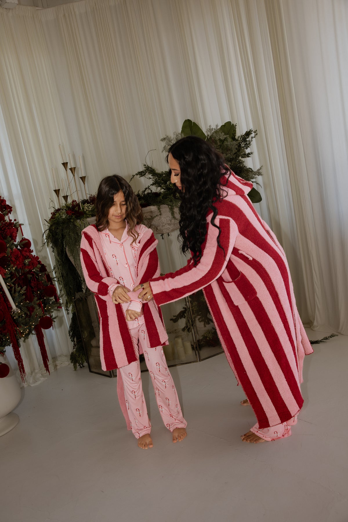 A woman helps a girl put on a pink and red striped Children’s Robe | Sugar Plum Stripe, both wearing matching candy cane print pajamas.