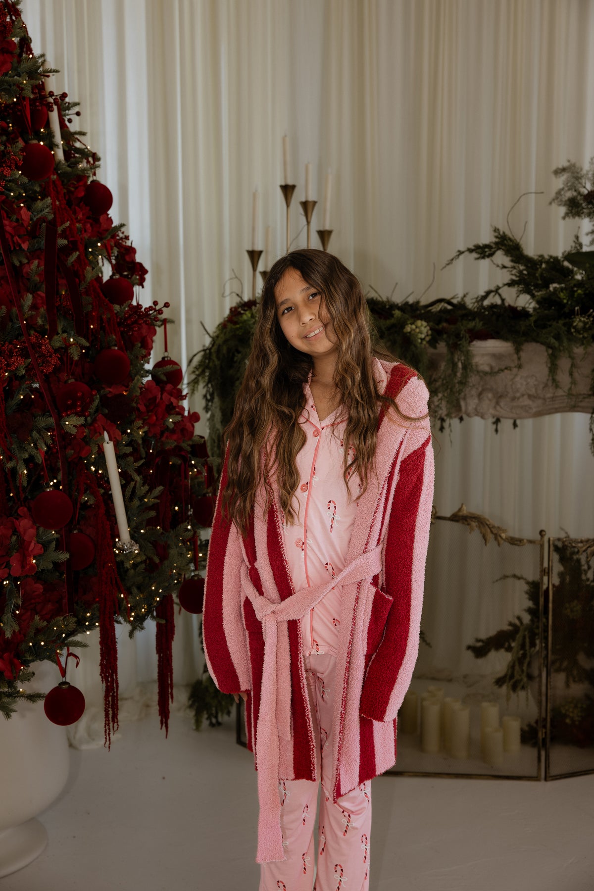 A girl wearing a pink candy cane print pajama set and a forever french baby Children’s Robe | Sugar Plum Stripe standing in front of a decorated Christmas tree.