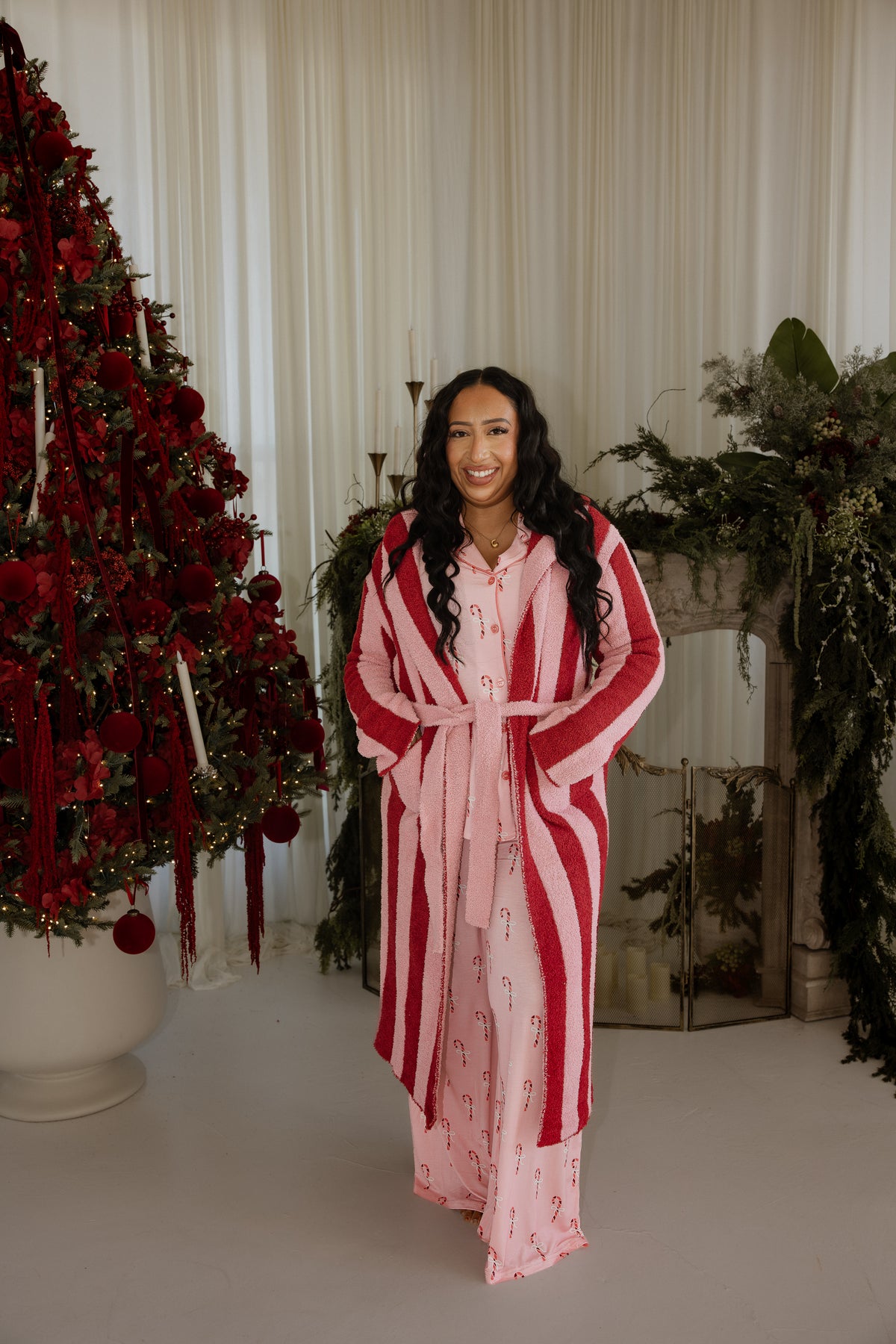 A smiling woman wearing a *PRE-ORDER* Adult Robe | Sugar Plum Stripe in pink and red striped microfiber feather yarn over matching candy-cane print pajamas, standing in front of a Christmas tree.