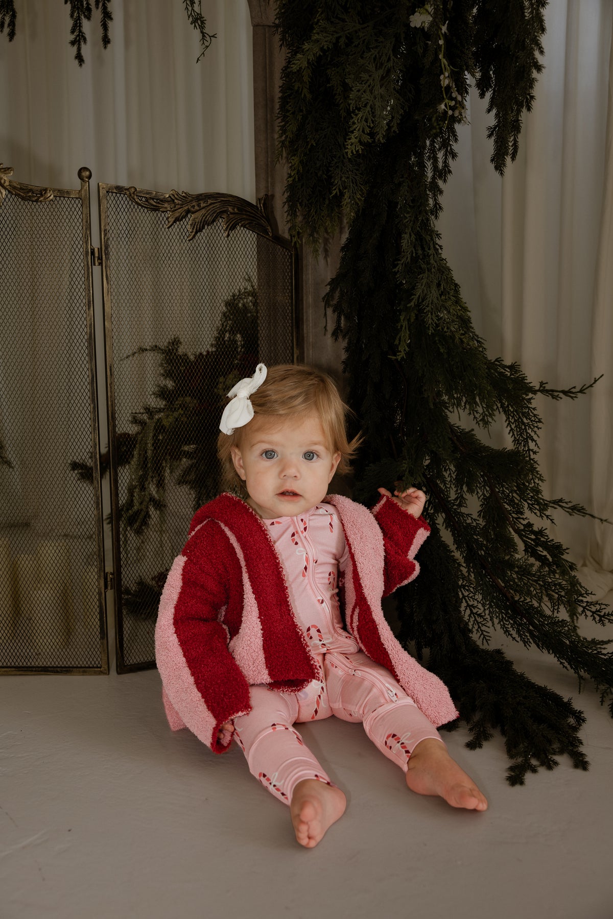 A toddler wears a Children’s Robe | Sugar Plum Stripe in color-blocked pink and red over a patterned pink onesie while sitting on the floor.
