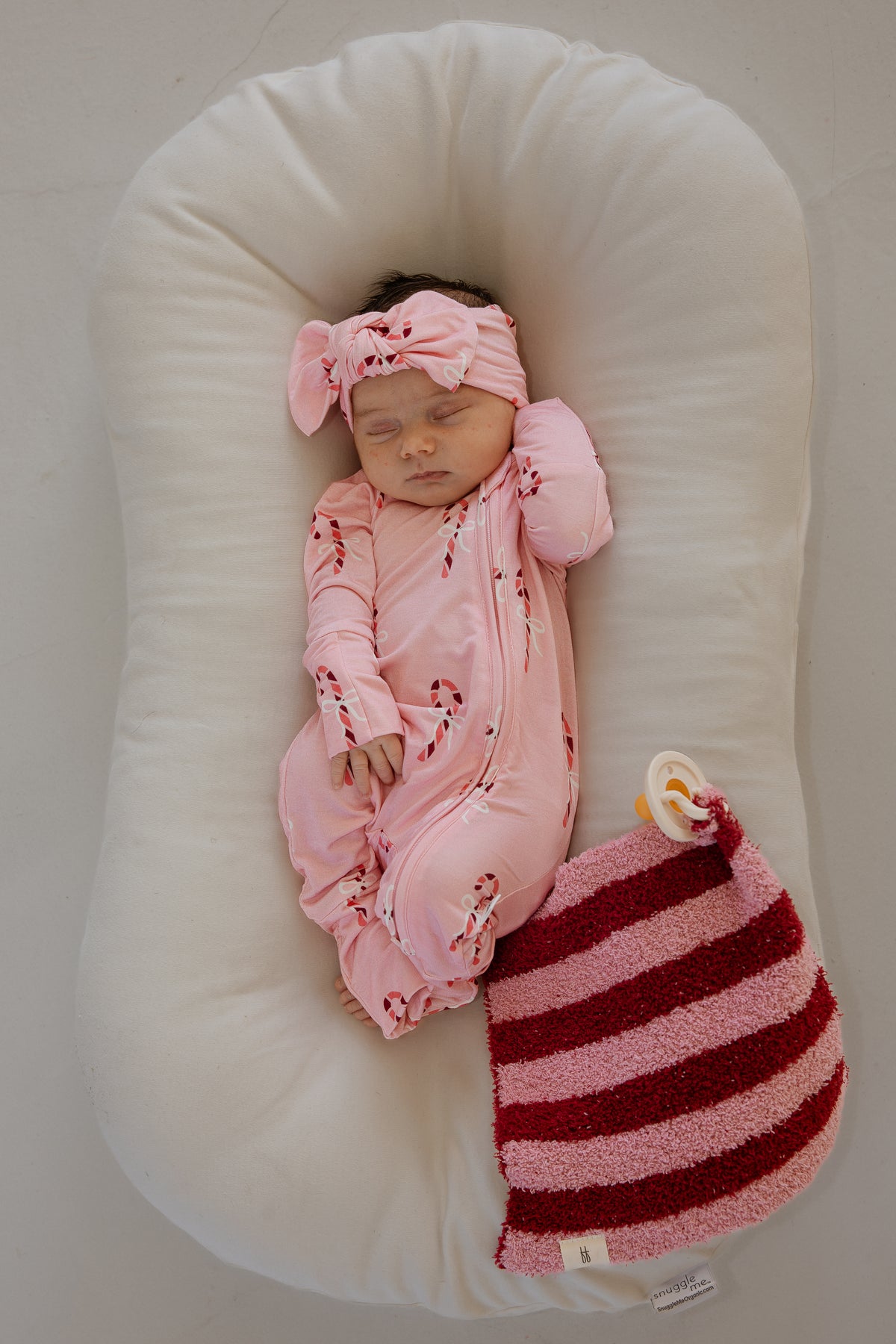 A sleeping newborn in a pink candy cane onesie and bow headband rests in a lounger next to a Sugar Plum Stripe lovey by forever french baby and a pacifier.