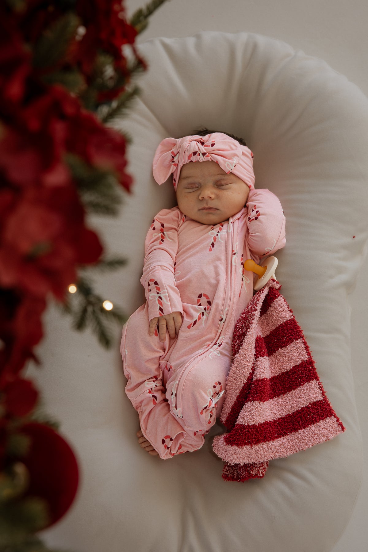 A sleeping infant wears forever french baby Bamboo Zip Pajamas | Pink Candy Cane with a matching knotted headband, resting next to a red and pink striped blanket and a pacifier.