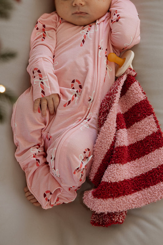 A baby in pink candy-cane print pajamas lies next to a Sugar Plum Stripe forever french baby lovey and a pacifier.