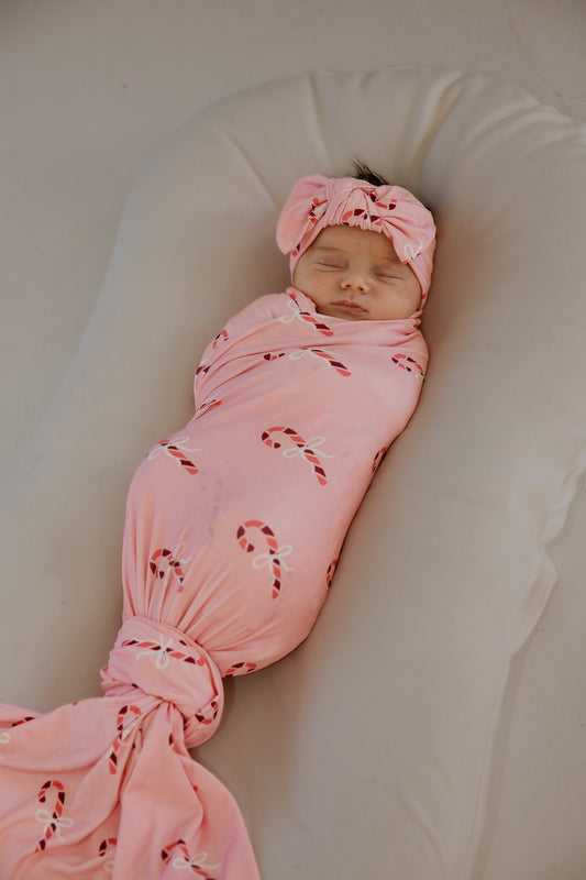 A newborn baby wearing a Bamboo Swaddle | Pink Candy Cane and matching headband, sleeping in a white lounger.