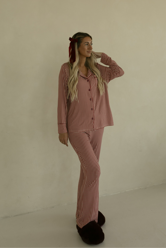 A woman with long blonde hair smiles to the side while wearing forever french baby's Long Sleeve Women's Bamboo Pajamas in Red Gingham, a soft, hypo-allergenic lounge set, paired with dark slippers against a plain beige wall.