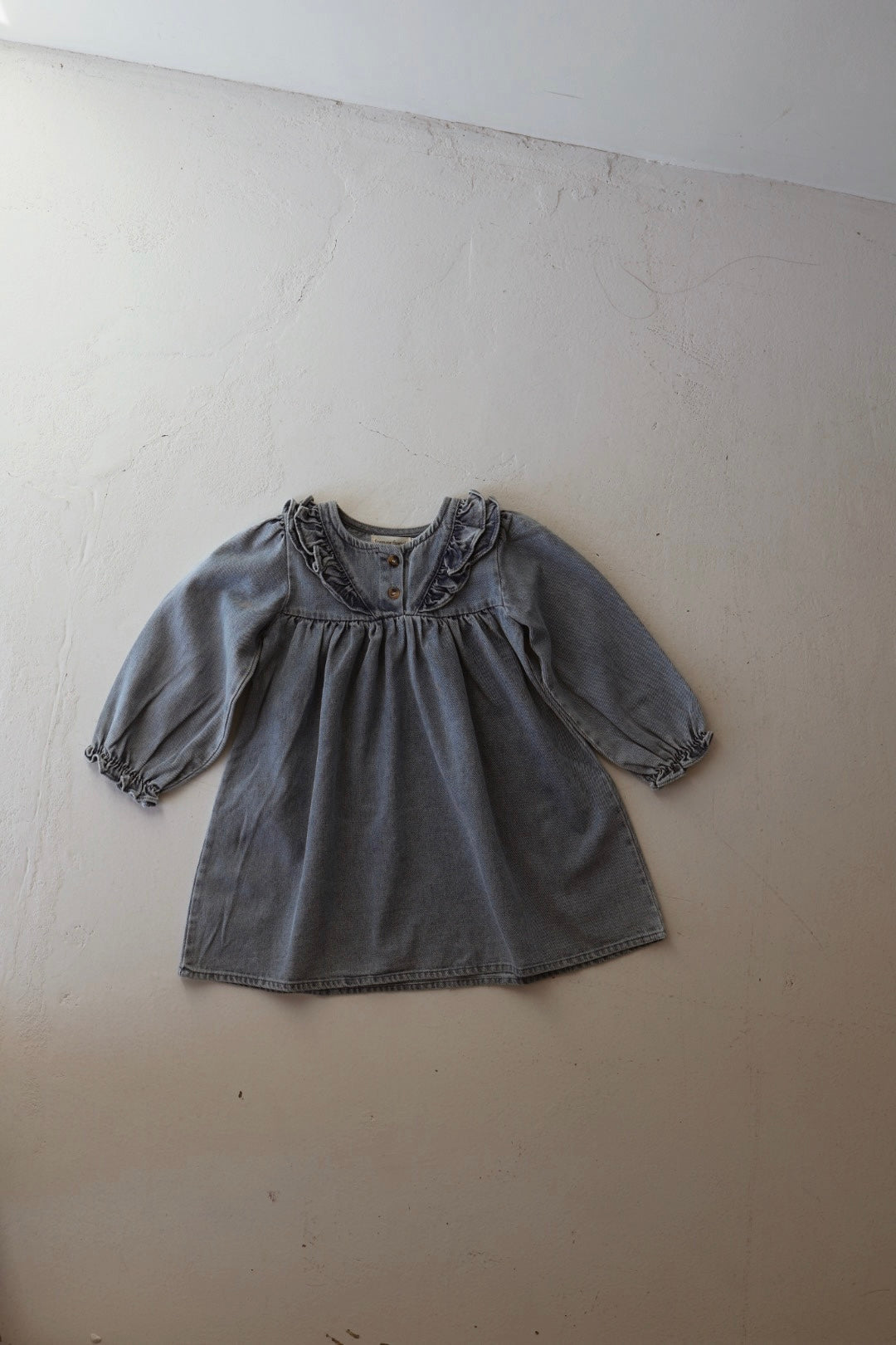 The forever french baby Denim Dress | Ruffle, a long-sleeved, light blue tunic with ruffled collar and cuffs, is laid flat on a light-colored textured surface.