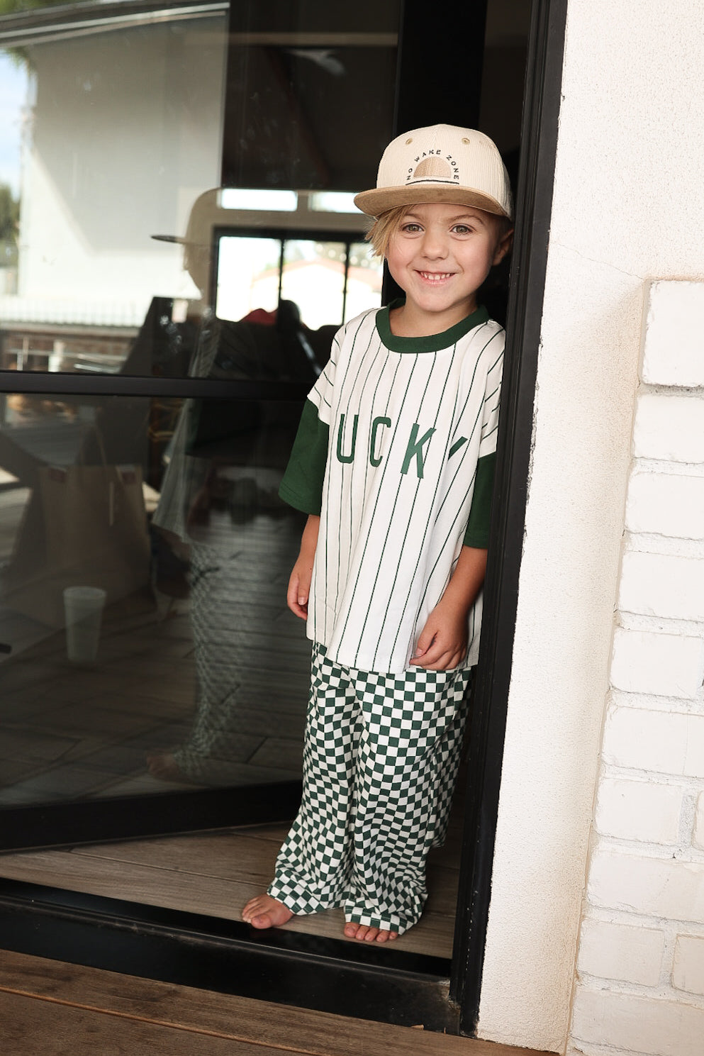 A young child in the forever french baby T-Shirt & Pant Set | Lucky Micro Check stands barefoot in a doorway, smiling at the camera.