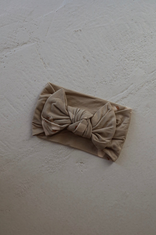 The Bamboo Head Wrap from forever french baby by Silly Goose, in beige with a large bow, rests on a textured light surface.