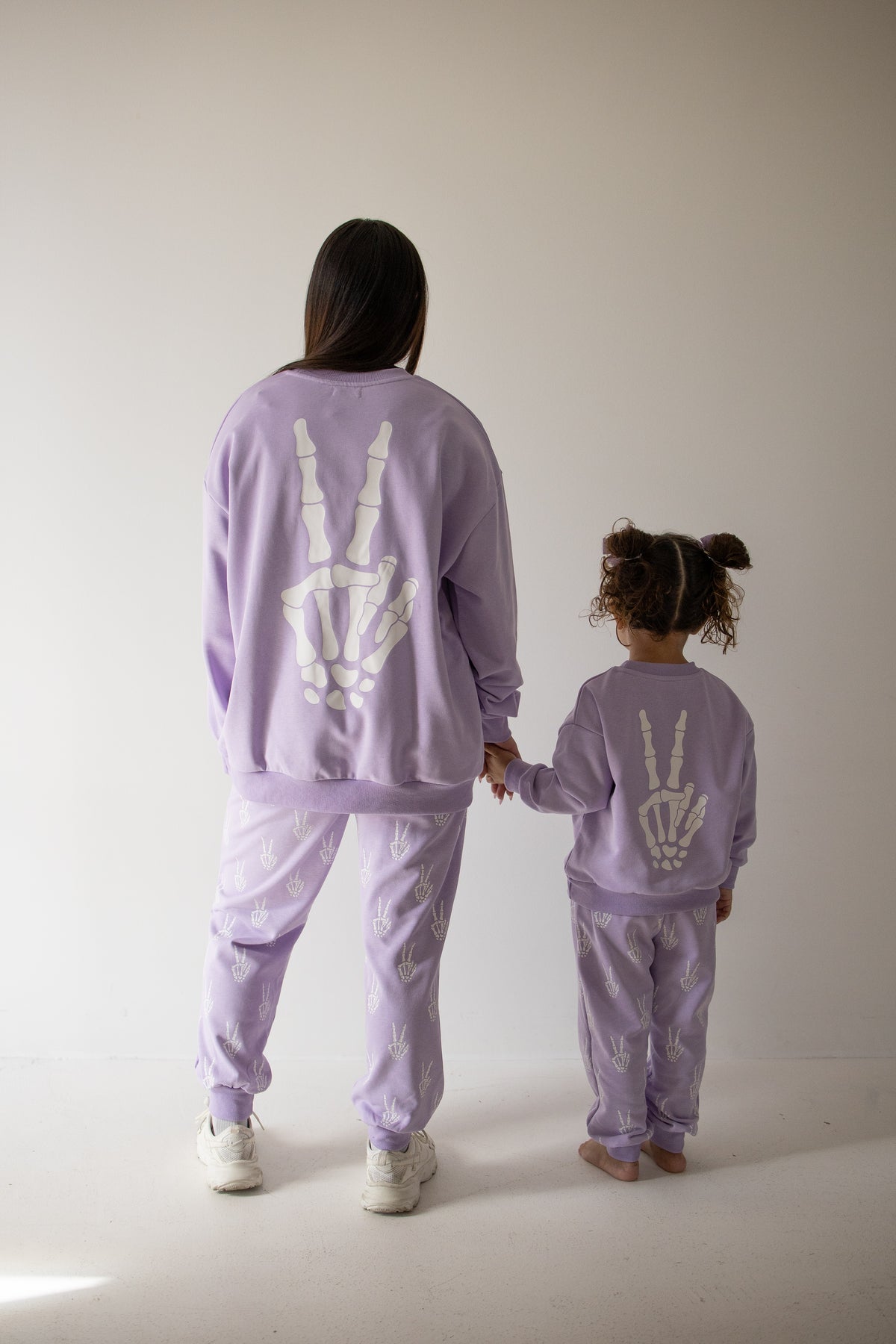 A woman and child, both dressed in matching forever french baby Women’s Sweat Set | Enchanted Skelly outfits featuring white skeleton hand designs on the back, stand hand in hand facing away from the camera against a simple light background—a fun and playful choice for families looking to coordinate their spooky season style.