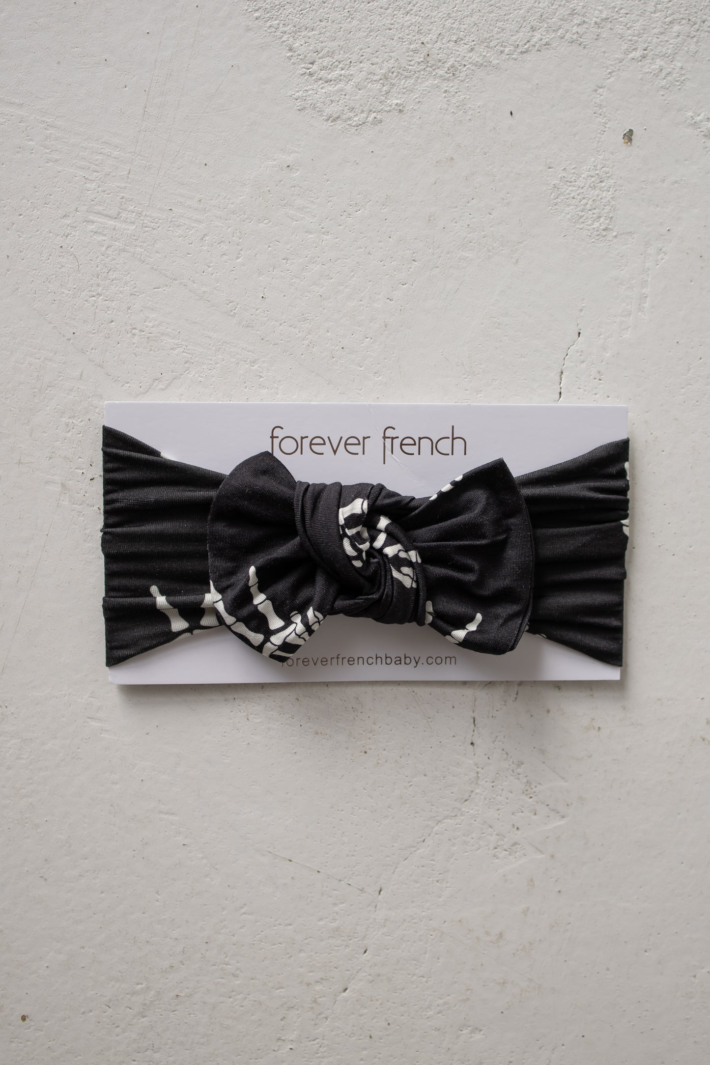 A “Midnight Skelly” bamboo head wrap by forever french baby, featuring a black knotted bow, displayed on a branded forever french card against a textured white background. This adjustable baby headband offers both comfort and style for your little one.
