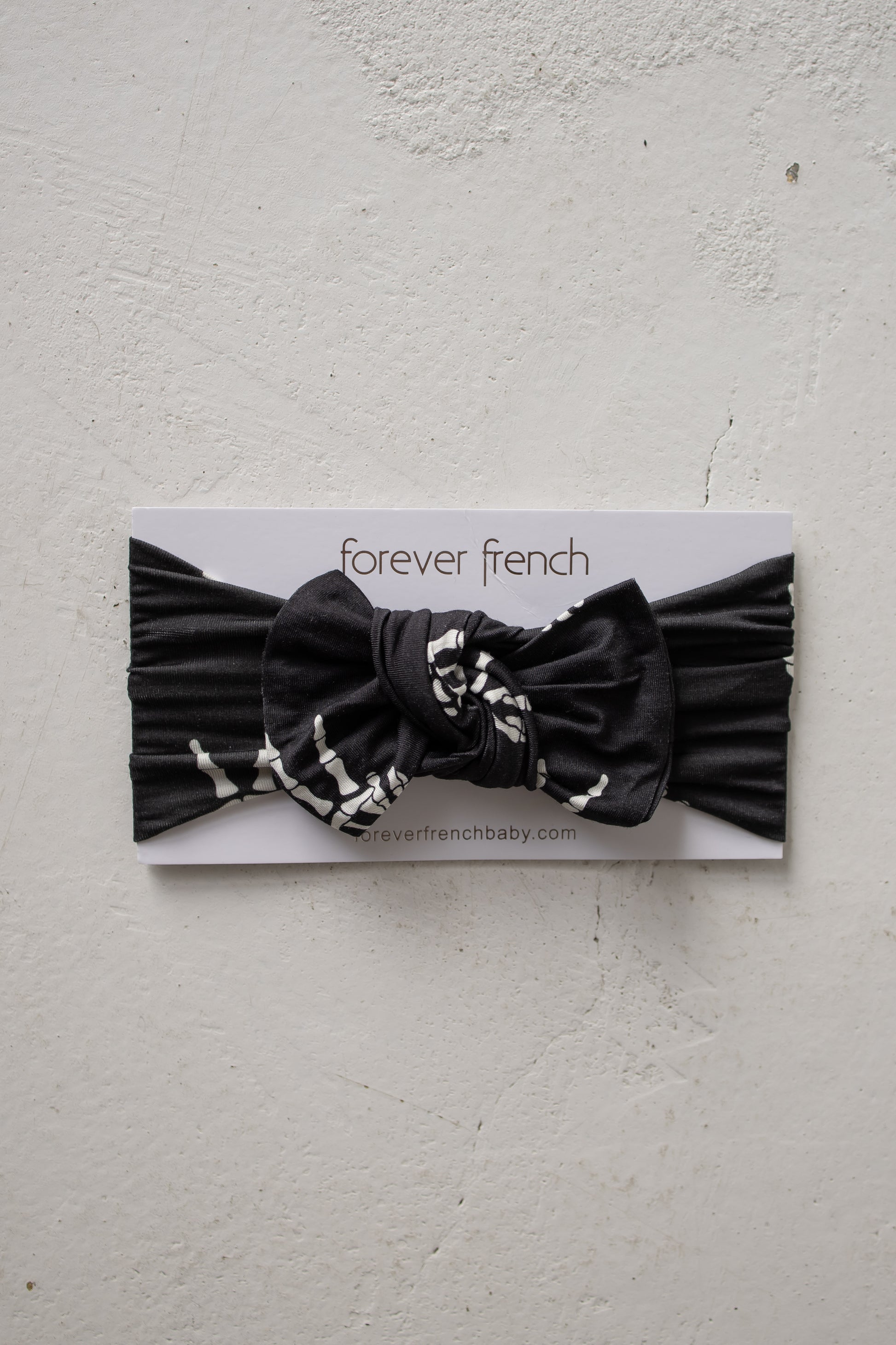 A “Midnight Skelly” bamboo head wrap by forever french baby, featuring a black knotted bow, displayed on a branded forever french card against a textured white background. This adjustable baby headband offers both comfort and style for your little one.