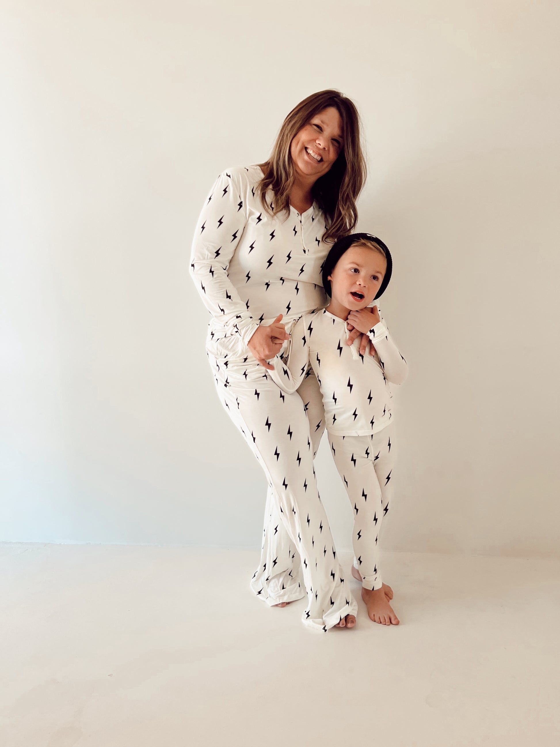 A woman with long, wavy hair and a young child stand against a plain background, both smiling and wearing matching Women's Bamboo Pajamas by forever french baby, featuring a white and black lightning bolt print. The barefoot child dons a black hat and strikes a playful pose. These pajamas are also breastfeeding-friendly.