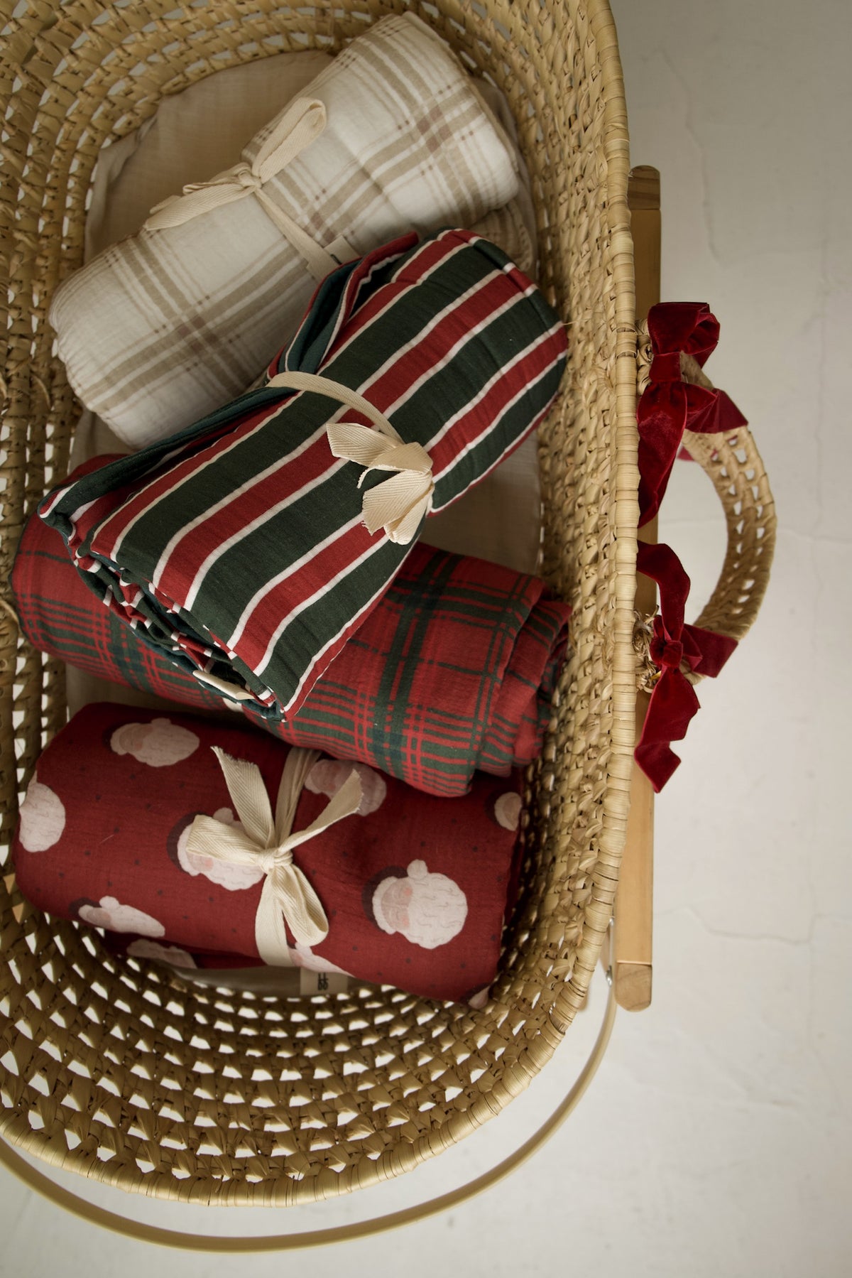 Three rolled blankets in a woven basket, featuring a Muslin Quilt | Christmas Plaid, a red Santa print, and a neutral plaid muslin cotton baby quilt.
