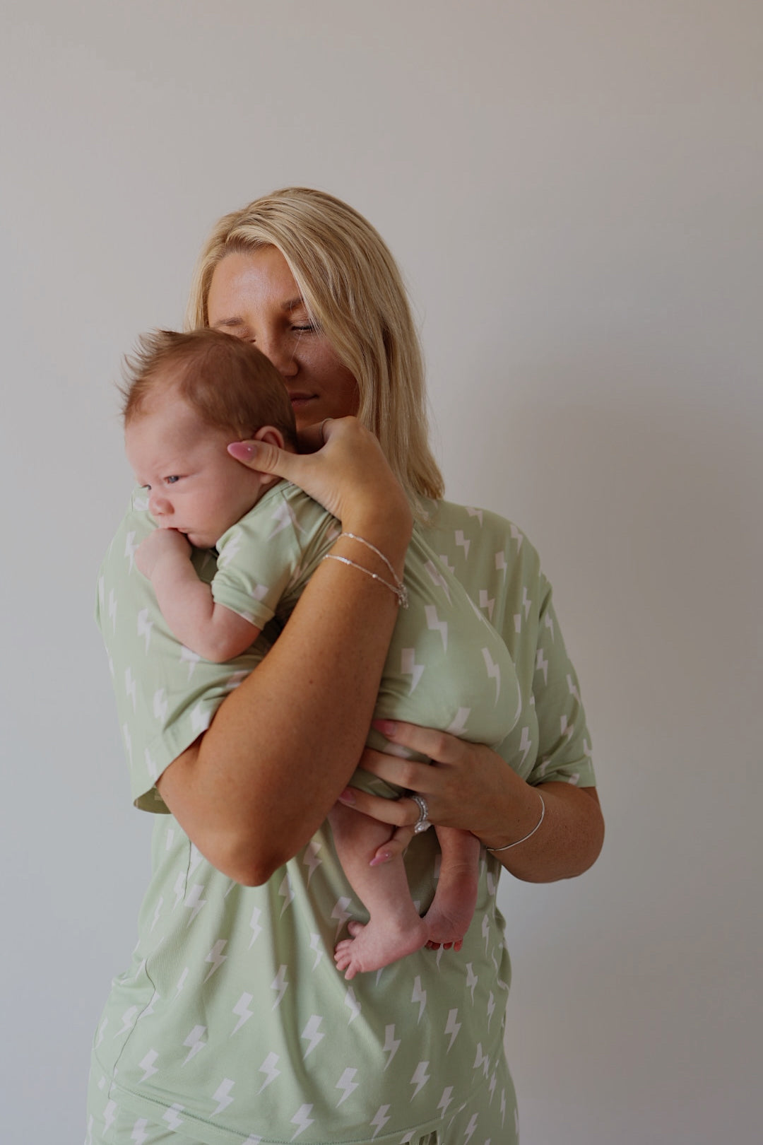 A blonde woman in a green lightning bolt-patterned outfit holds a newborn dressed in forever french baby’s Shortie Bamboo Zip | Flash pajamas, perfectly matching her. The background is plain and light-colored.