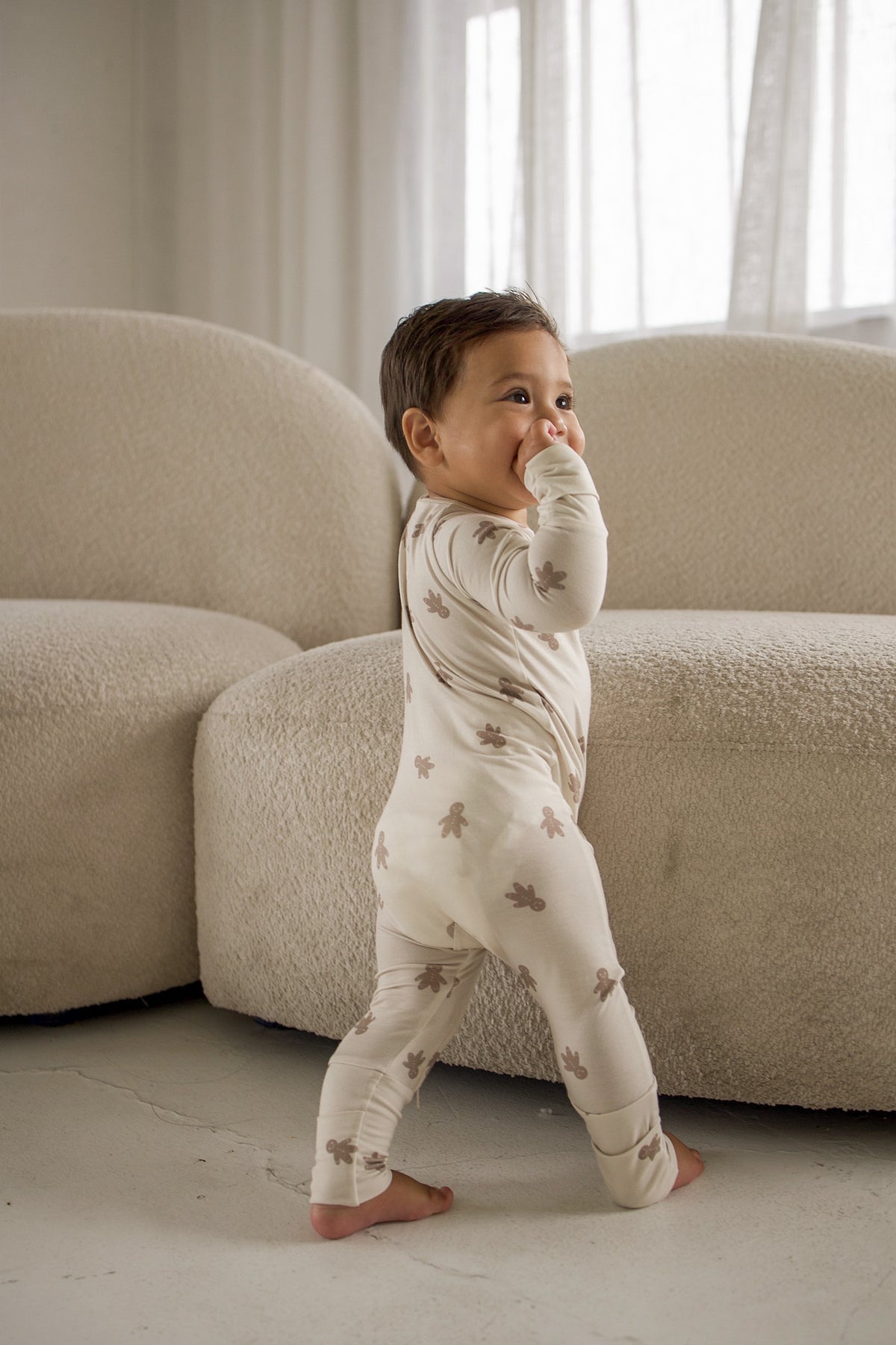 A toddler wearing cream-colored Bamboo Zip Pajamas | Gingersnap, featuring a zip-up sleeper pattern of small gingerbread men.
