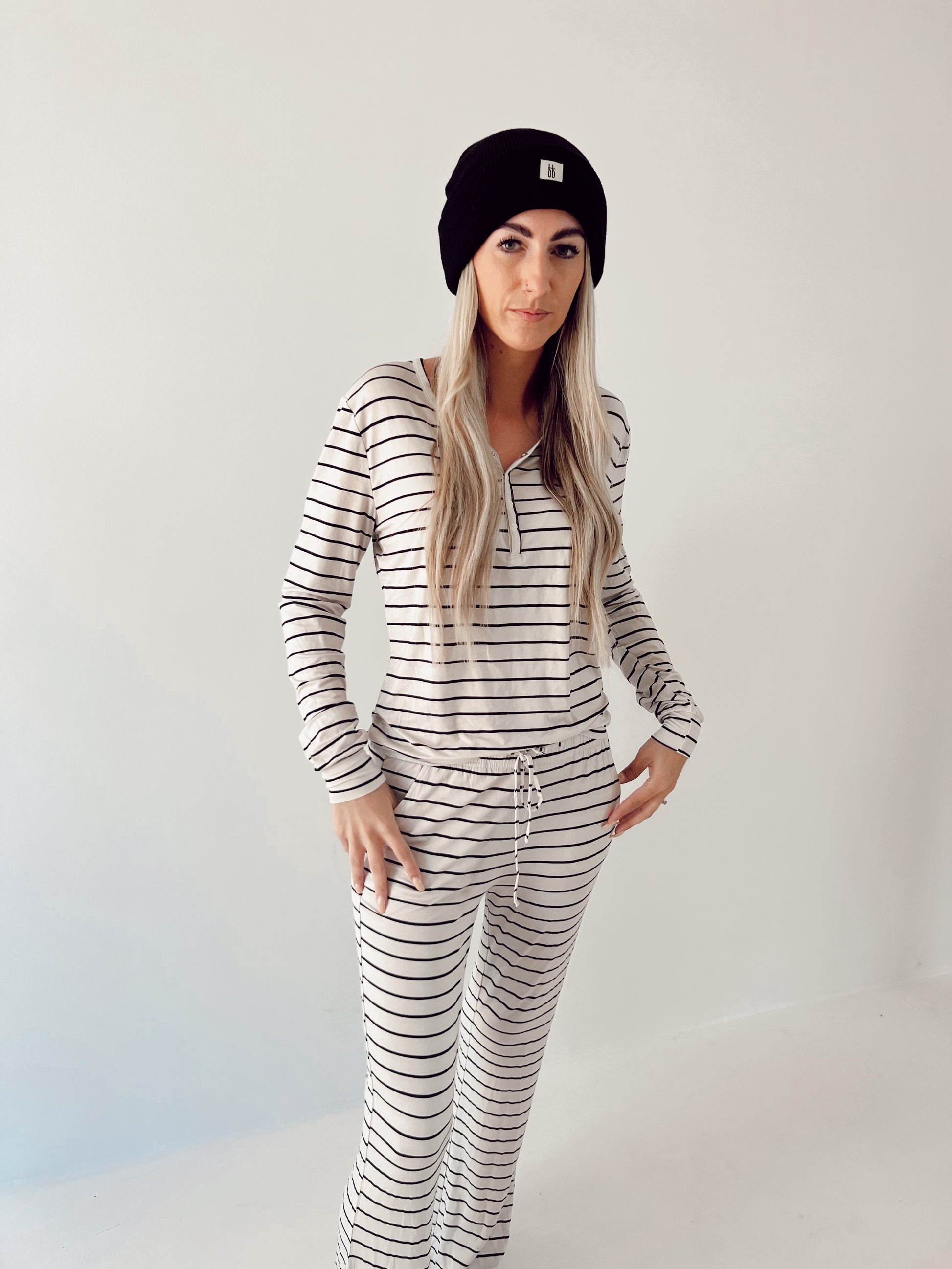 Wearing a black beanie, a long-haired person stands against a plain light background in "forever french baby" Women’s Bamboo Pajamas | Grey & Black Stripe. The breathable set includes horizontal striped, long-sleeved top and pants.