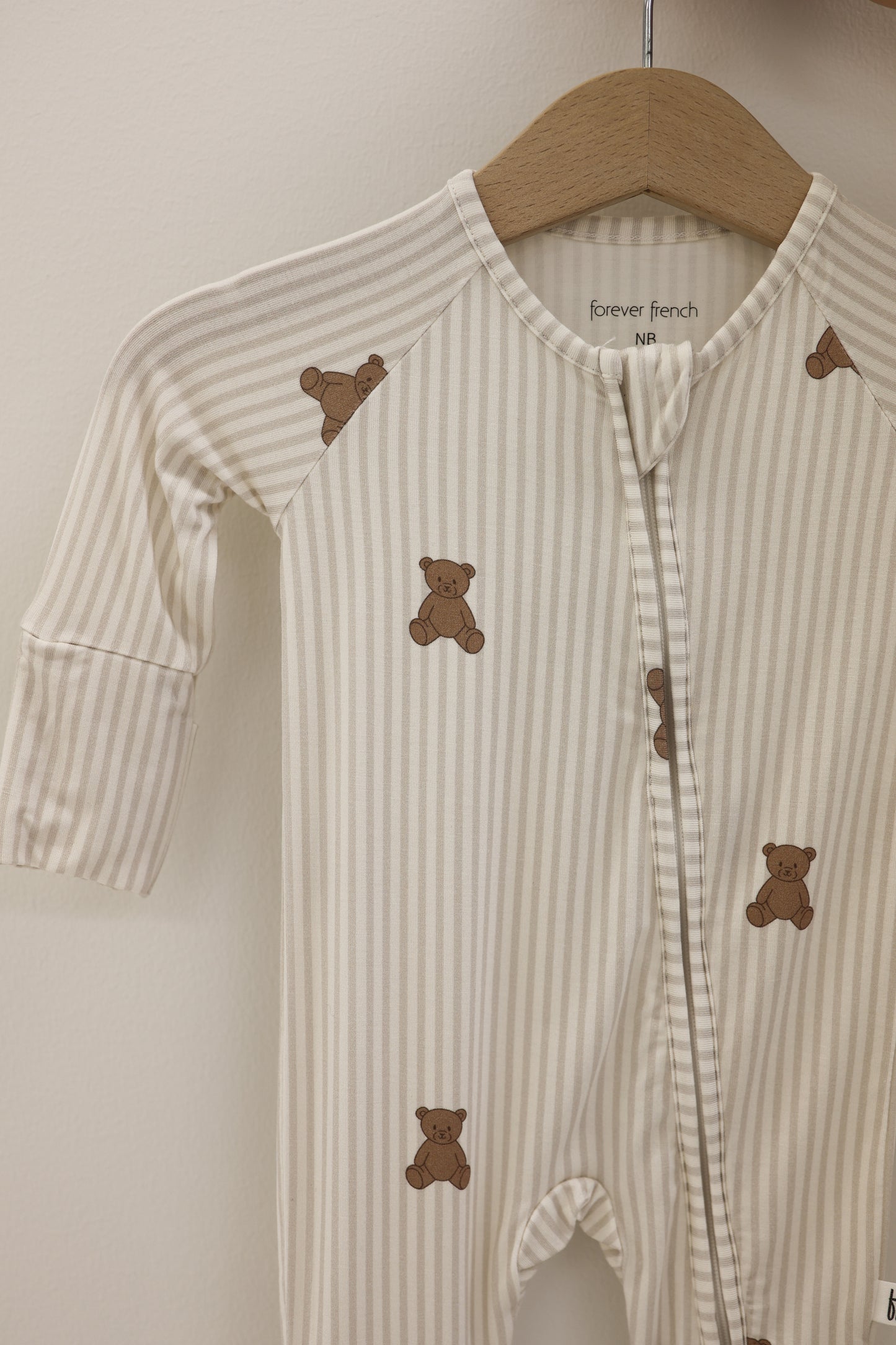 The Bamboo Zip Pajamas | Teddy by forever french baby, featuring beige and white stripes with brown teddy bear prints, is displayed on a wooden hanger against a light wall—ideal for those seeking hypoallergenic baby clothing.
