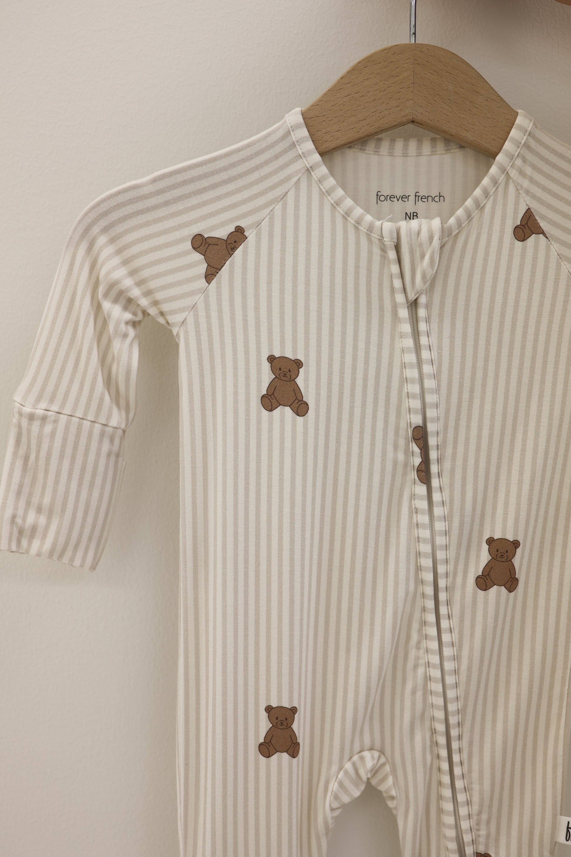 The Bamboo Zip Pajamas | Teddy by forever french baby, featuring beige and white stripes with brown teddy bear prints, is displayed on a wooden hanger against a light wall—ideal for those seeking hypoallergenic baby clothing.