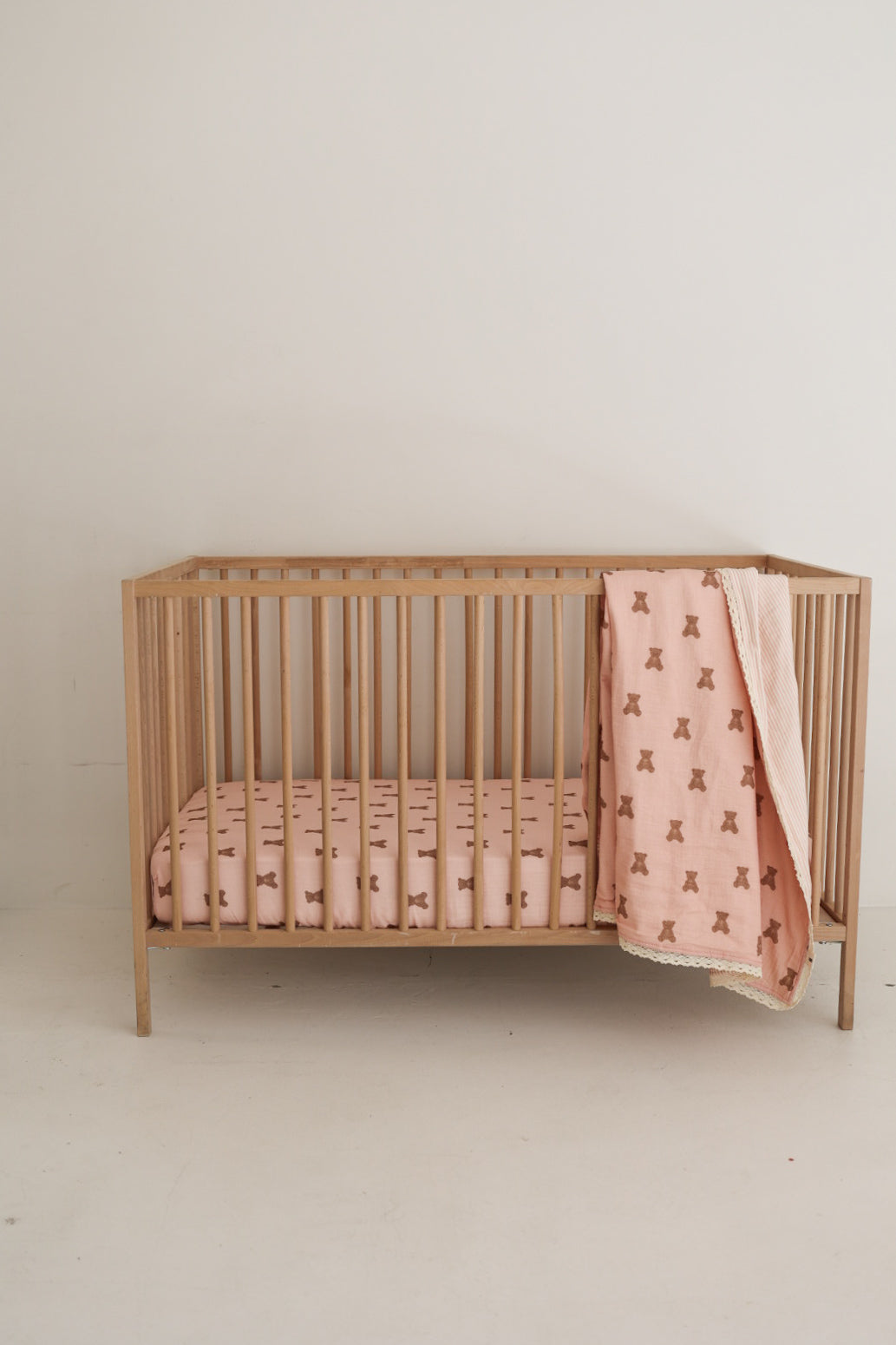 The Muslin Crib Sheet | Pink Lottie by forever french baby is on a wooden crib with a matching pink blanket featuring a brown pattern, creating cozy bedding against a plain white wall and floor.