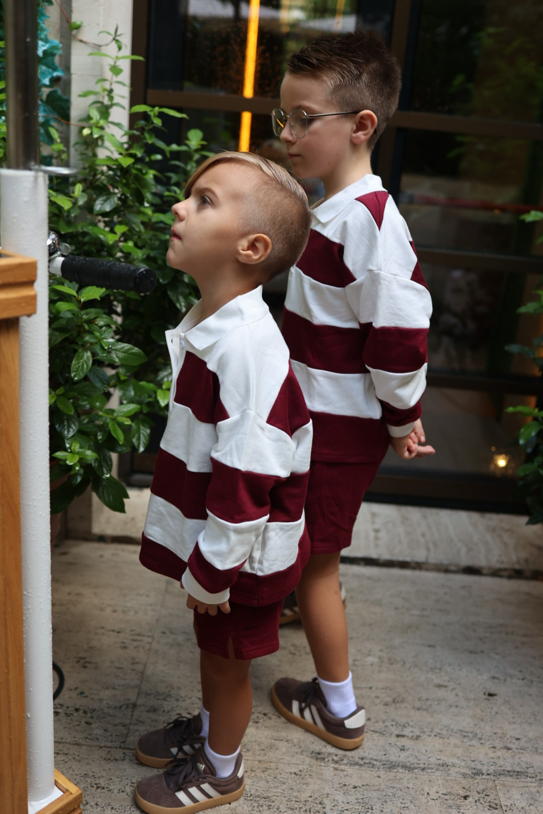 Two young boys wearing matching burgundy and white striped long-sleeved polo shirts and Child Terry Shorts in Deep Cranberry.