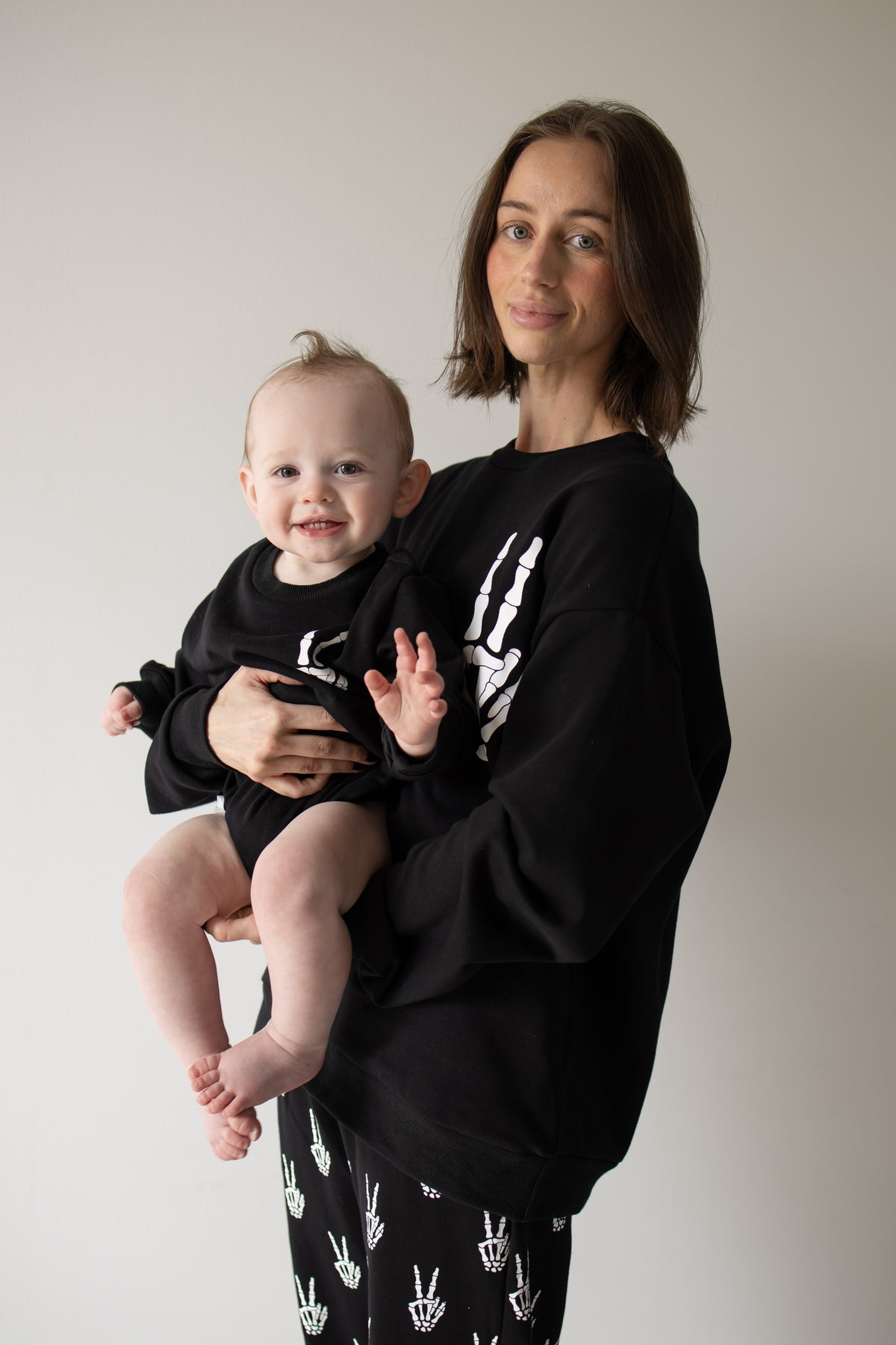 A woman holds a smiling baby, both wearing the forever french baby Women’s Sweat Set | Midnight Skelly, featuring matching black outfits with white skeleton hand designs, standing in front of a plain light background—perfect for spooky season.