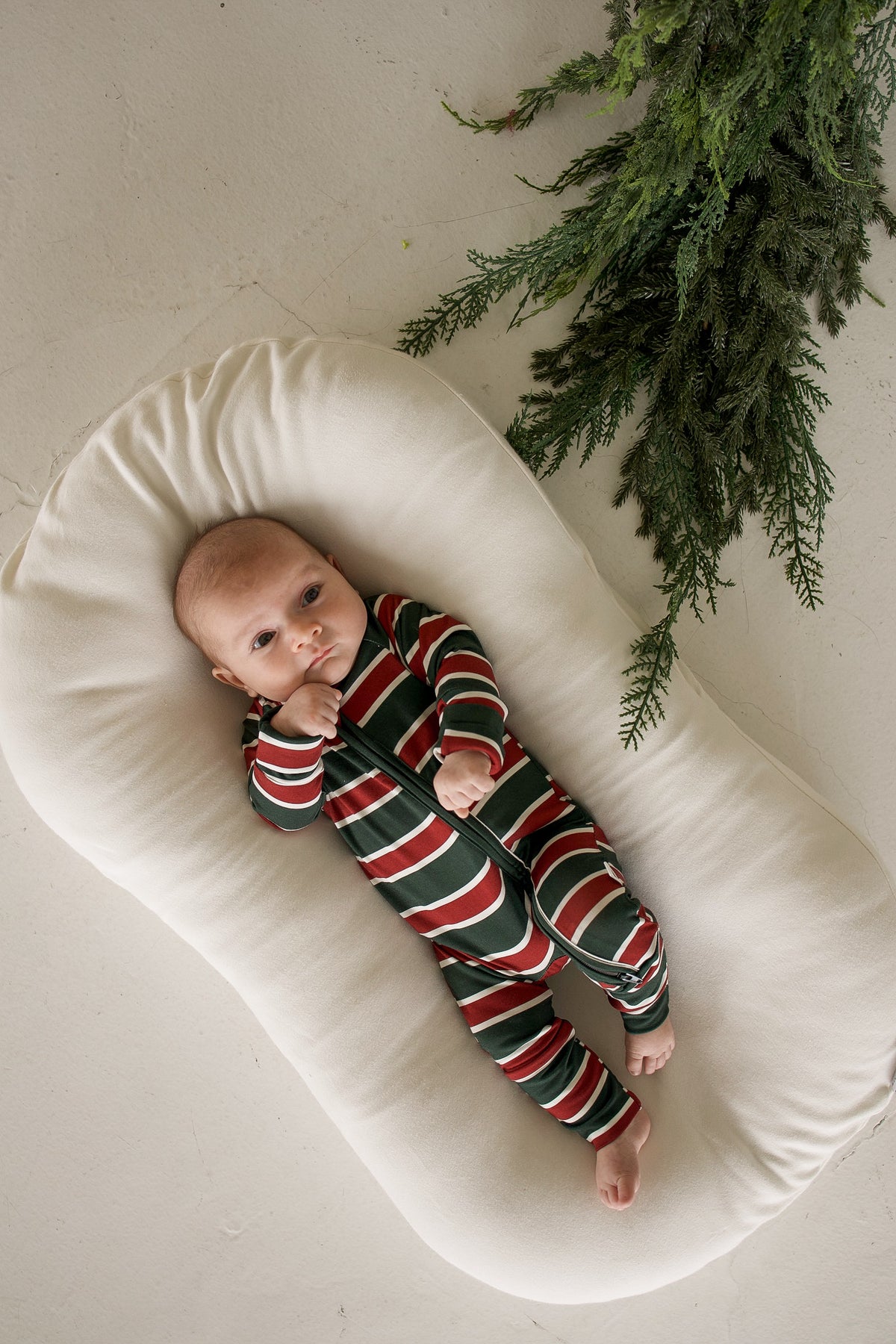 A baby wearing forever french baby Bamboo Zip Pajamas in Holiday Stripe while lying on a cream-colored lounger.