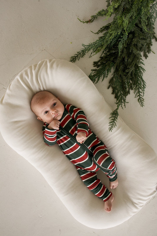A baby wearing forever french baby Bamboo Zip Pajamas in Holiday Stripe while lying on a cream-colored lounger.