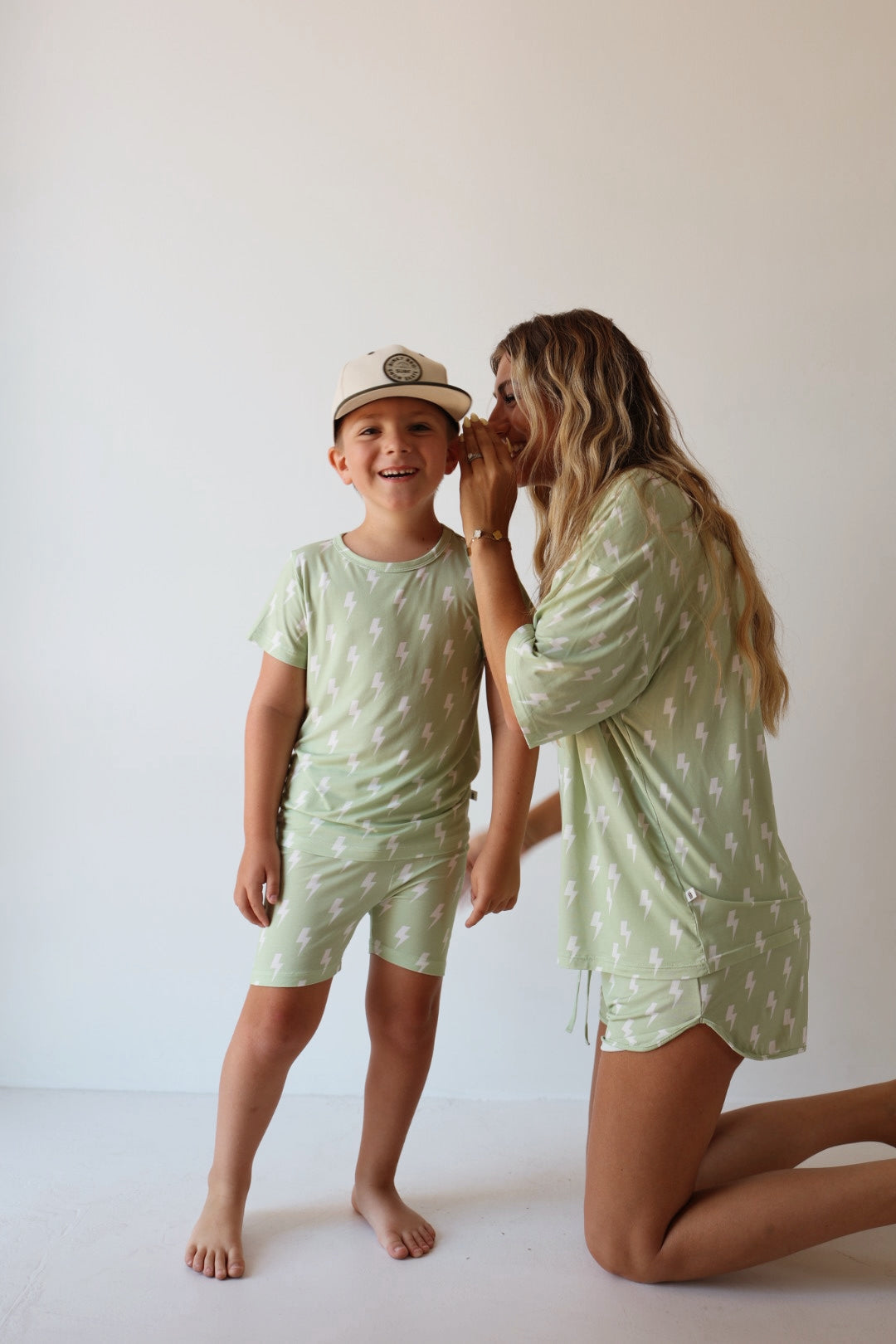A woman kneels beside a smiling young boy, whispering in his ear. Both wear forever french baby Bamboo Short Set | Flash—light green pajamas with white lightning bolts. The boy also wears a beige cap against a plain, light background.