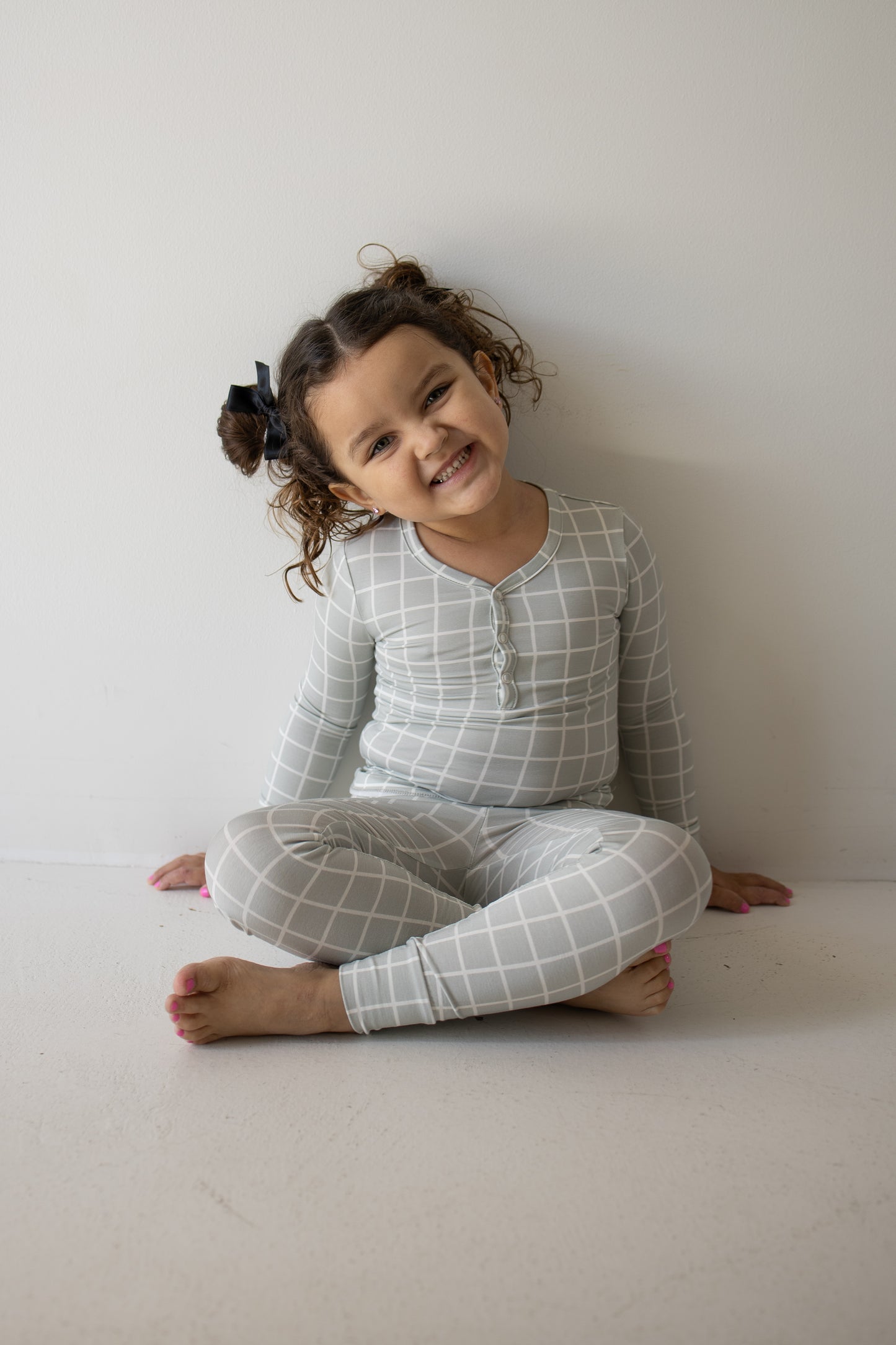 A young child with curly hair in pigtails, dressed in the forever french baby Bamboo Two Piece Pajamas in the Foggy Grid pattern, sits cross-legged on the floor against a white wall, smiling widely.