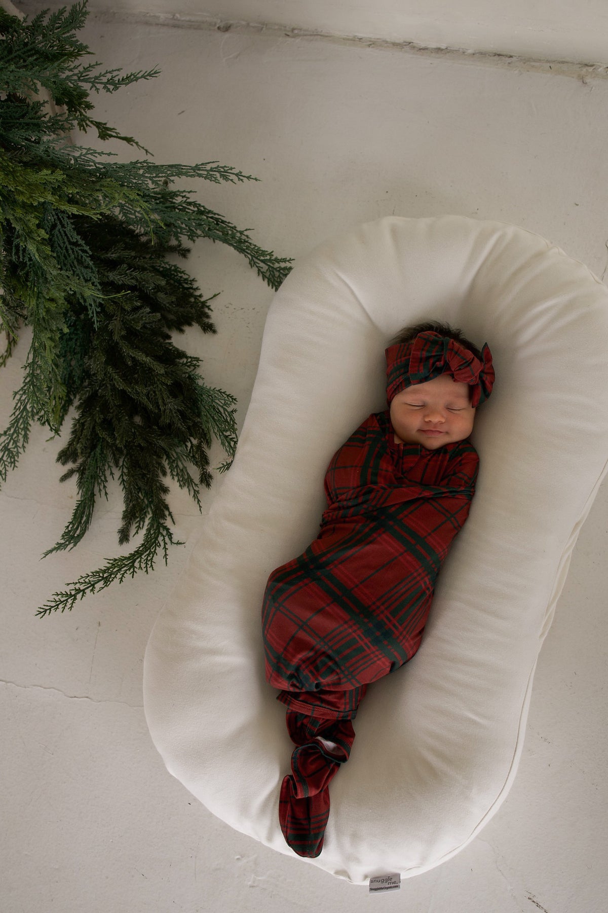 A newborn baby swaddled in a Bamboo Swaddle | Christmas Plaid and a matching knotted headband, resting in a white baby lounger.