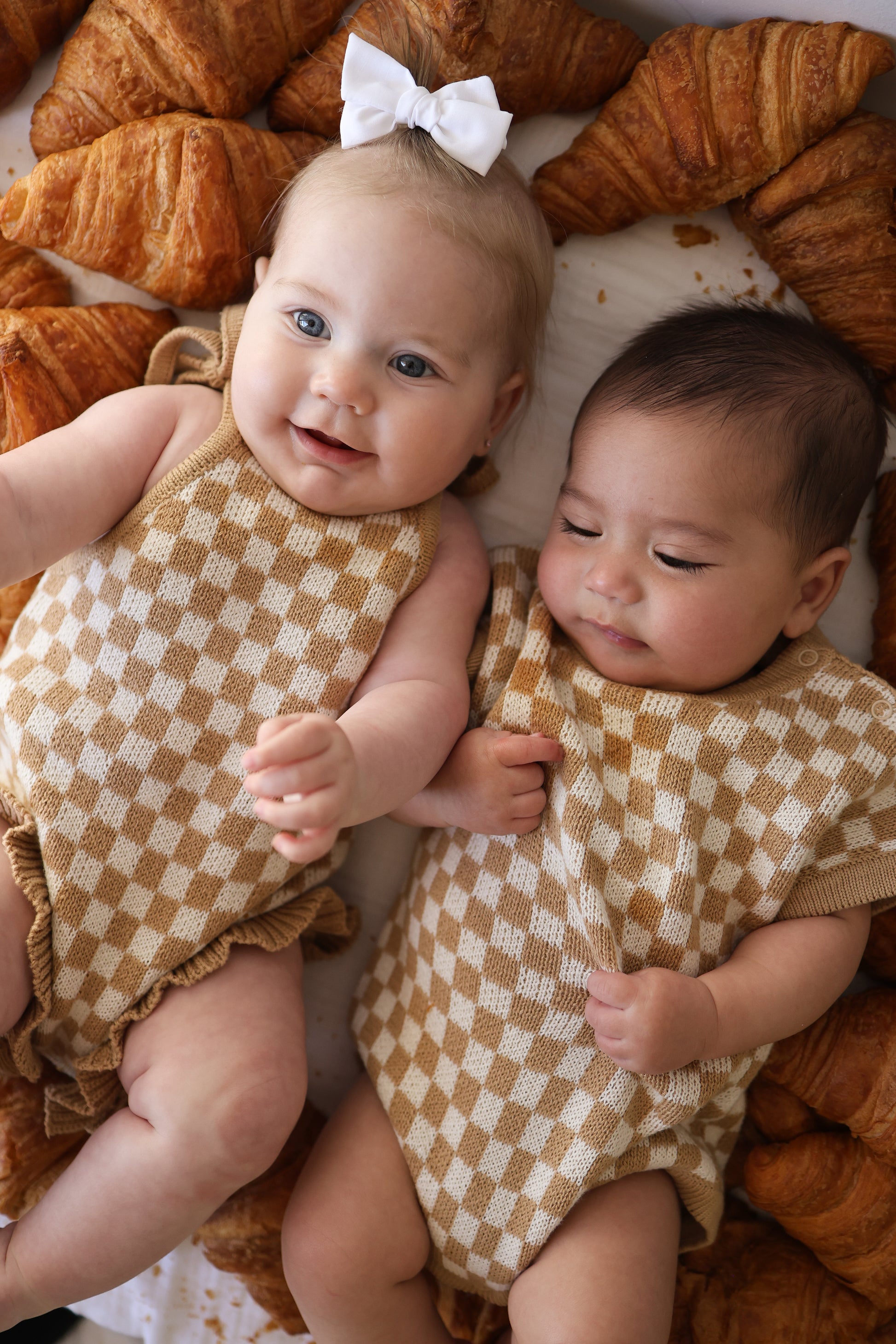 Two babies in matching forever french baby Knit Ruffle Romper | Latte Check lie side by side on a white blanket surrounded by croissants. One smiles with a white bow, while the other looks content and relaxed.