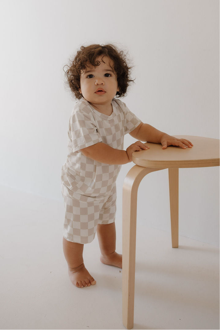 A toddler with curly hair wears the forever french baby Bamboo Short Set | Checkerboard in beige and white, standing by a light wooden table in a minimal, bright room.
