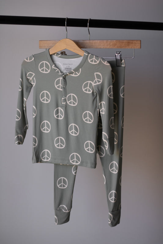 The "Bamboo Two Piece Pajamas | Peace" by forever french baby, featuring long sleeves and green fabric with white peace signs, hangs on a wooden hanger against a plain gray wall.