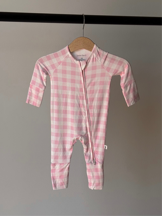 The forever french baby Bamboo Zip Pajamas | Pink Sugar features a pink and white checkered design, long sleeves and legs, a diagonal zipper from neck to leg, and is made from breathable bamboo fabric. Displayed on a hanger against a gray wall.